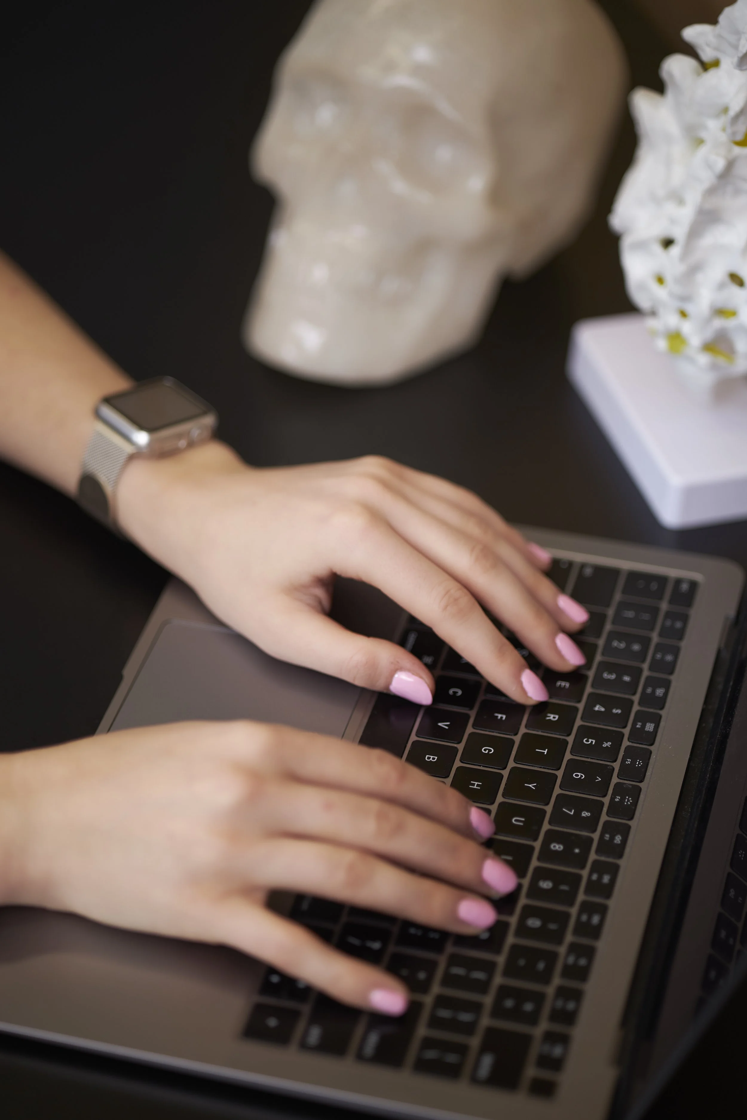 Person typing on a laptop keyboard with a skull sculpture and white flowers in the background.