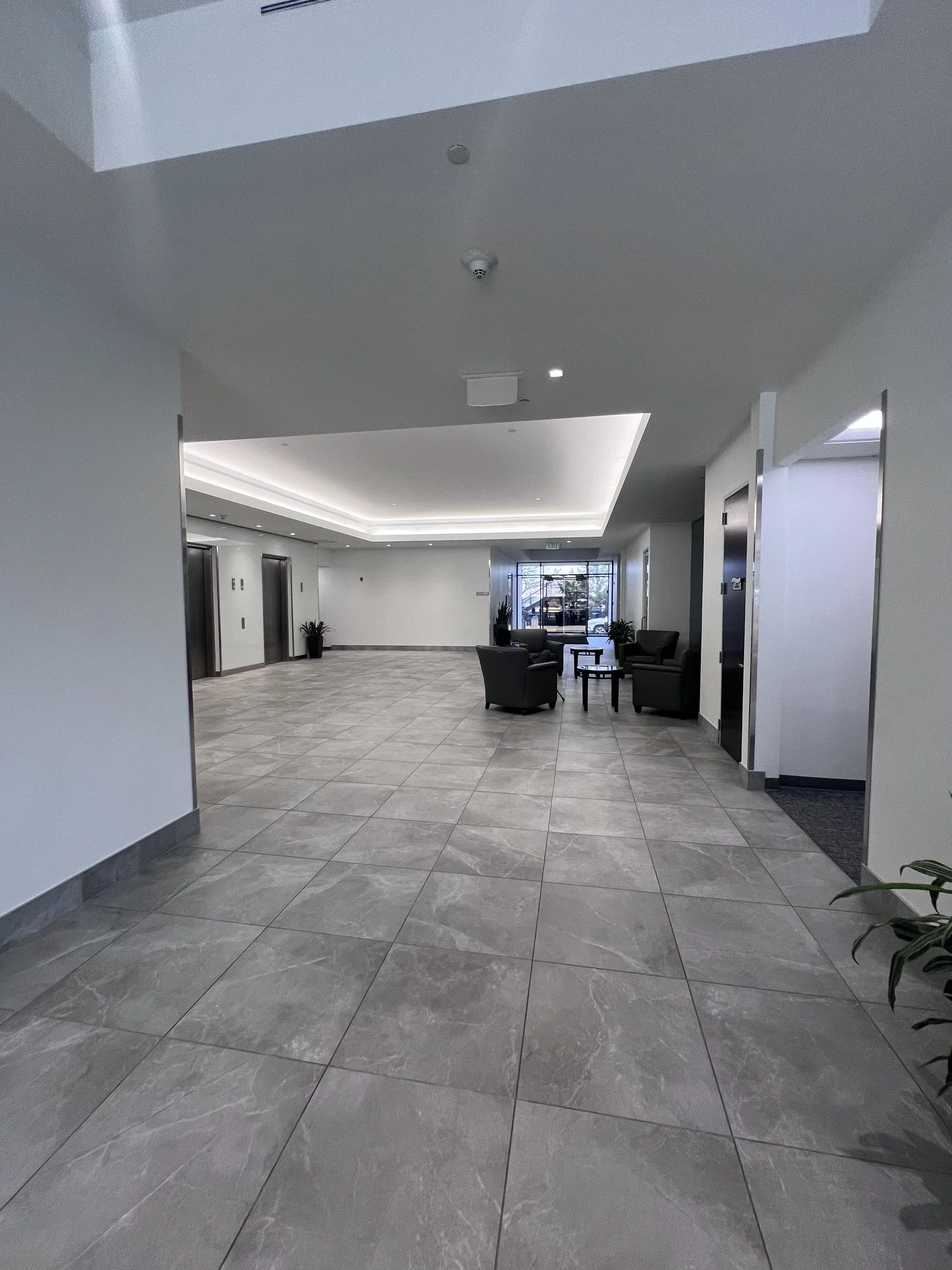 Interior of a modern building lobby with gray tiled floor, white walls, and a seating area with four black chairs and small round tables. There are potted plants near the elevators and a glass door at the entrance.