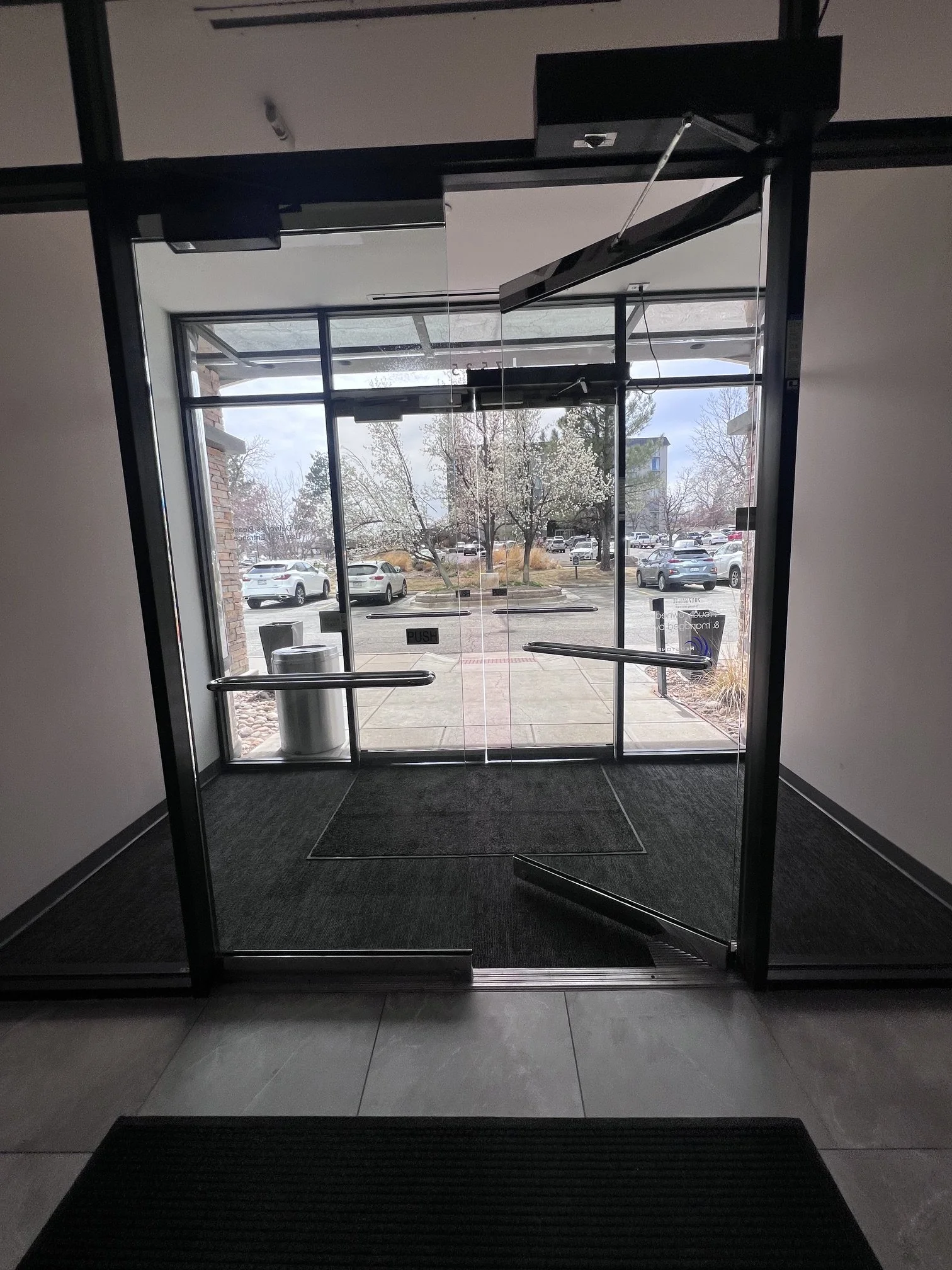 Glass door from inside looking out to a parking lot with trees and cars, through a large glass window. The door has black frames and push bars, with signs instructing to push or pull.
