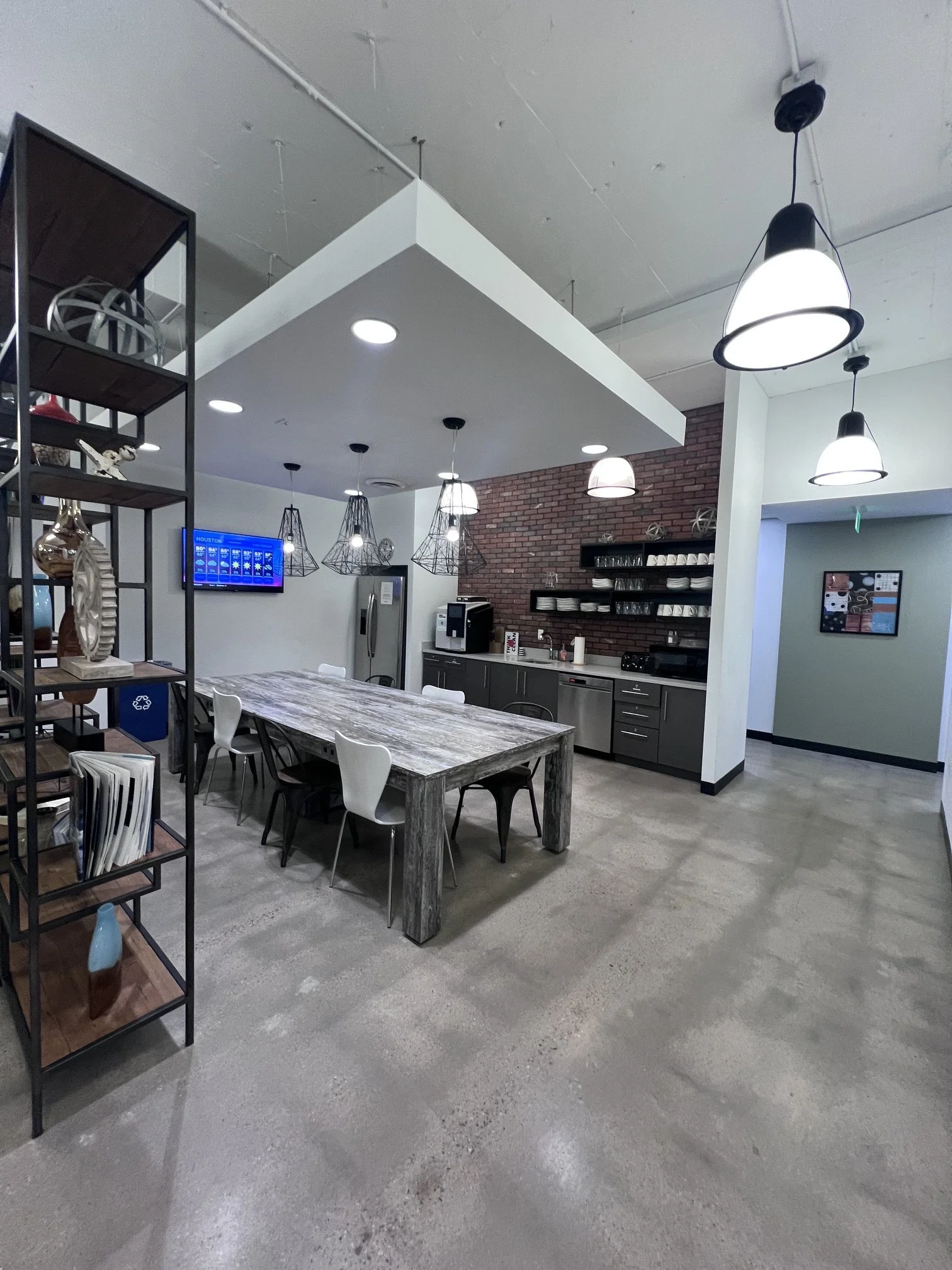 Interior of a modern kitchen and dining area with a concrete floor, wooden dining table, white chairs, and industrial-style pendant lights. There is a brick accent wall with shelves holding dishes and glassware, a refrigerator, microwave, and framed poster on the wall.