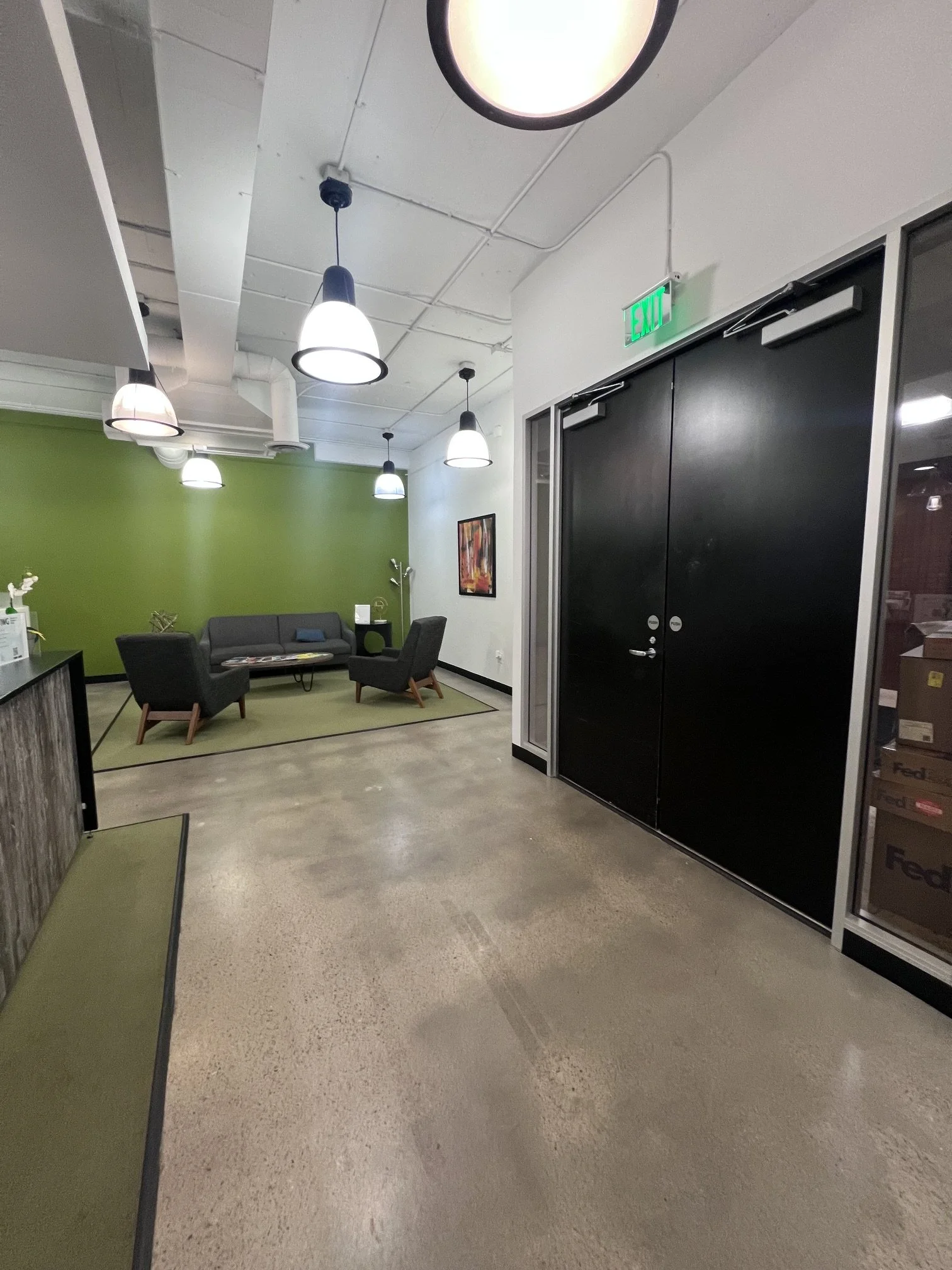 An interior space with a green accent wall, a sofa, several chairs, and hanging ceiling lights in an office or waiting area. There is a doorway with an illuminated green exit sign above it and a framed artwork on the wall.