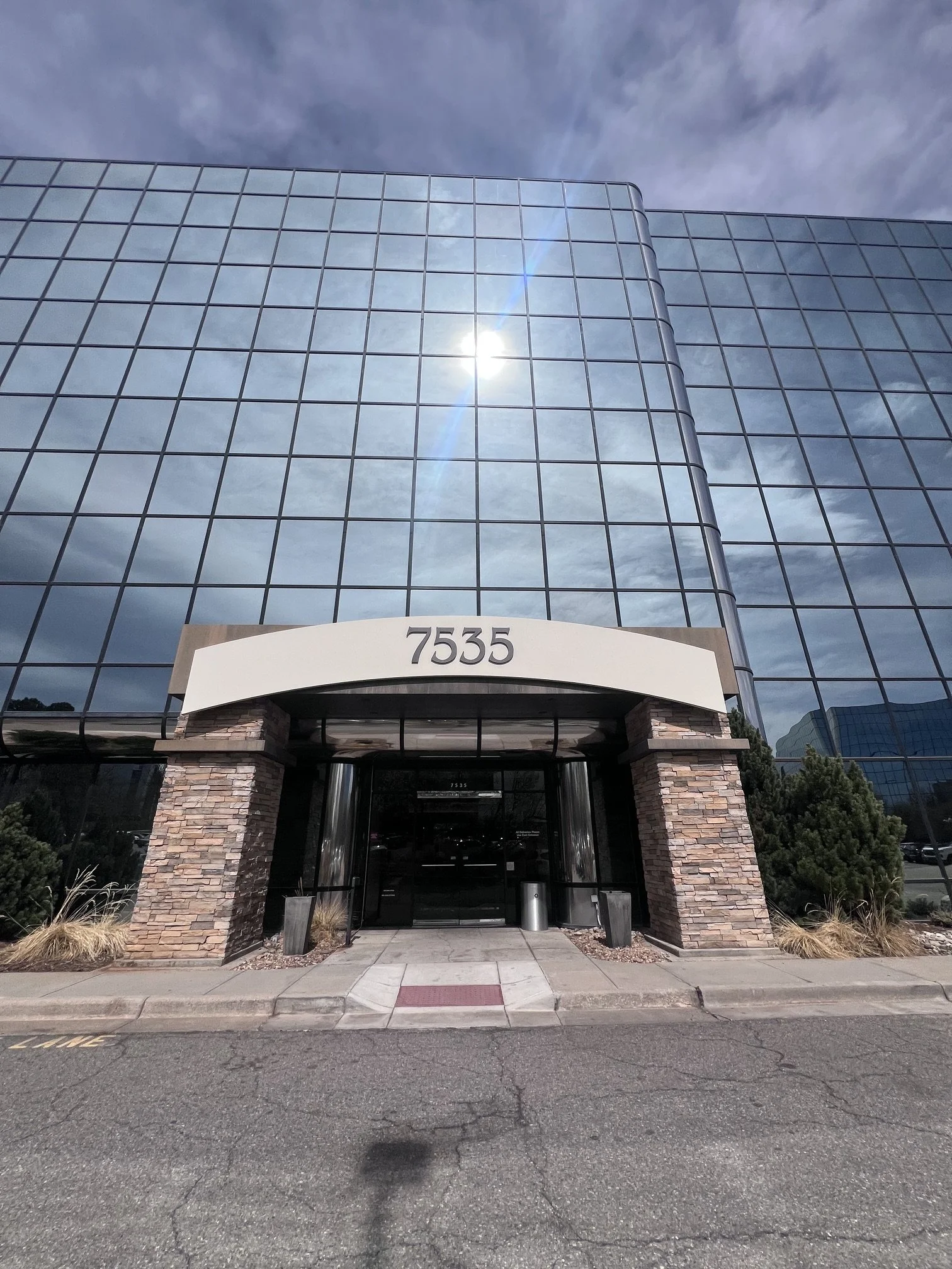 A modern glass office building with a curved entrance and stone accents, displaying the number 7535 above the door, under a partly cloudy sky with the sun reflecting off the windows.