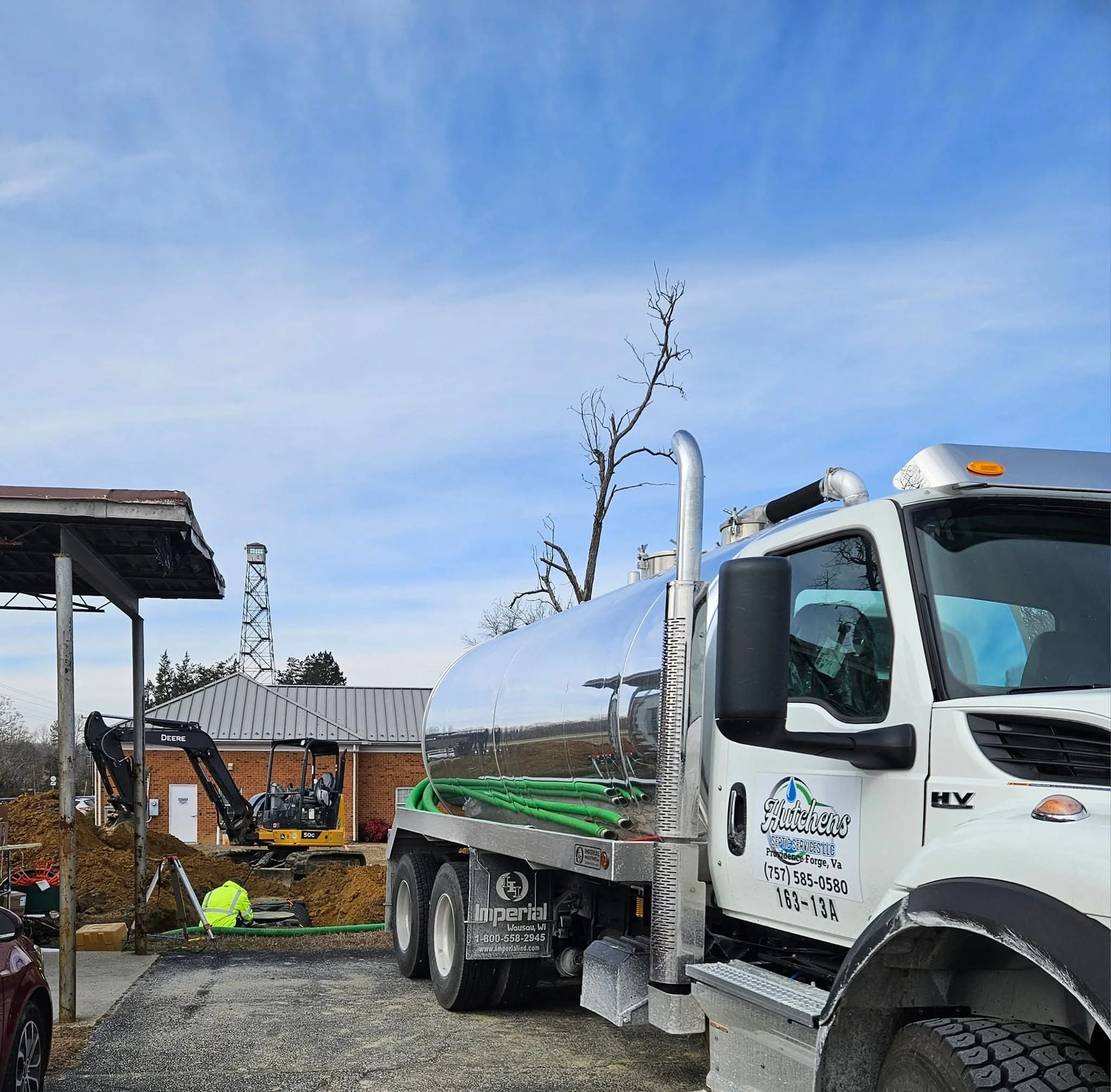 A septic pump truck is parked at a construction site with a worker and excavator in the background.