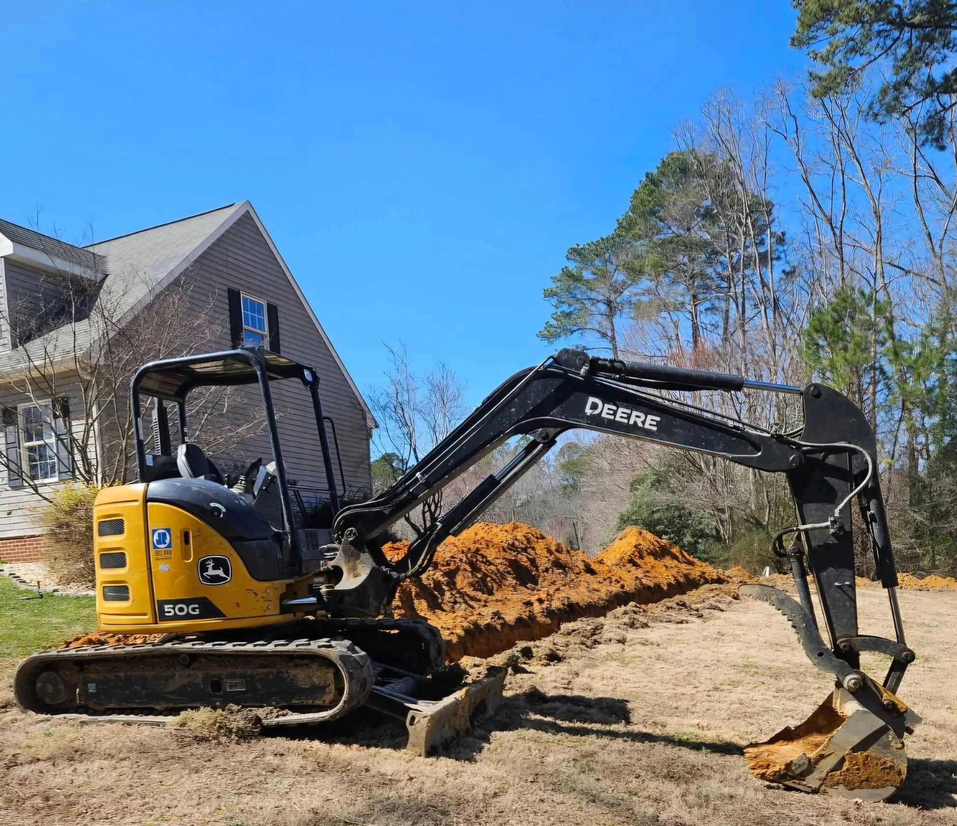 A John Deere excavator digging a trench in a yard near a multi-story house, with trees in the background under a bright blue sky.