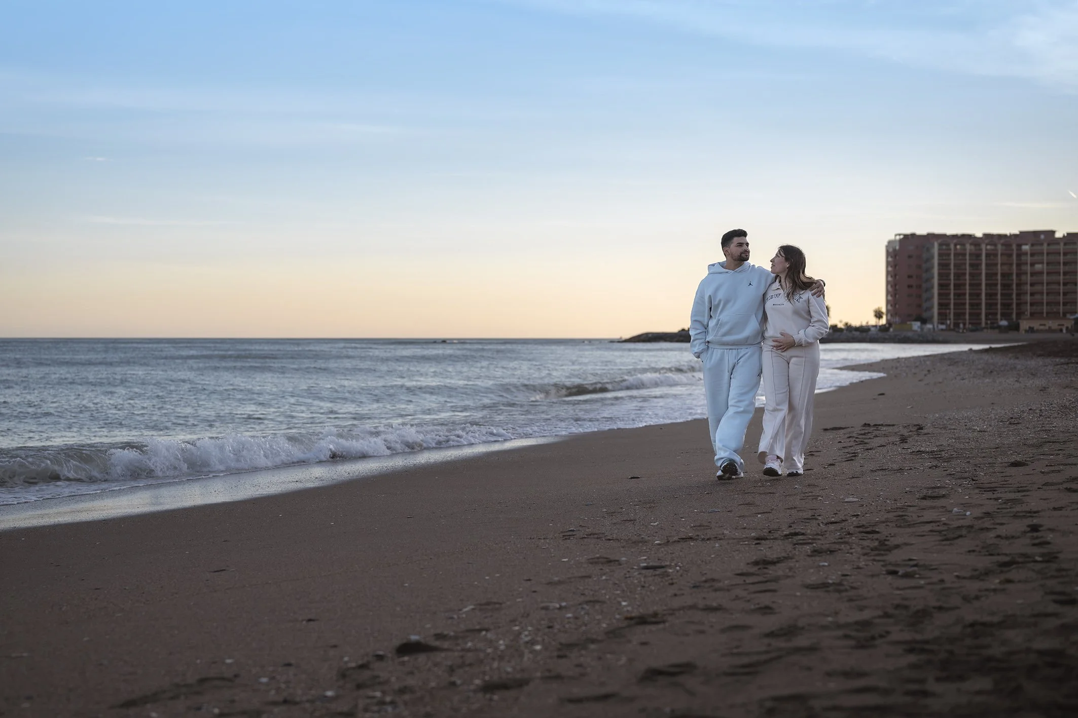 Una pareja caminando por la playa al atardecer, con un edificio alto en el fondo y el mar a su lado.