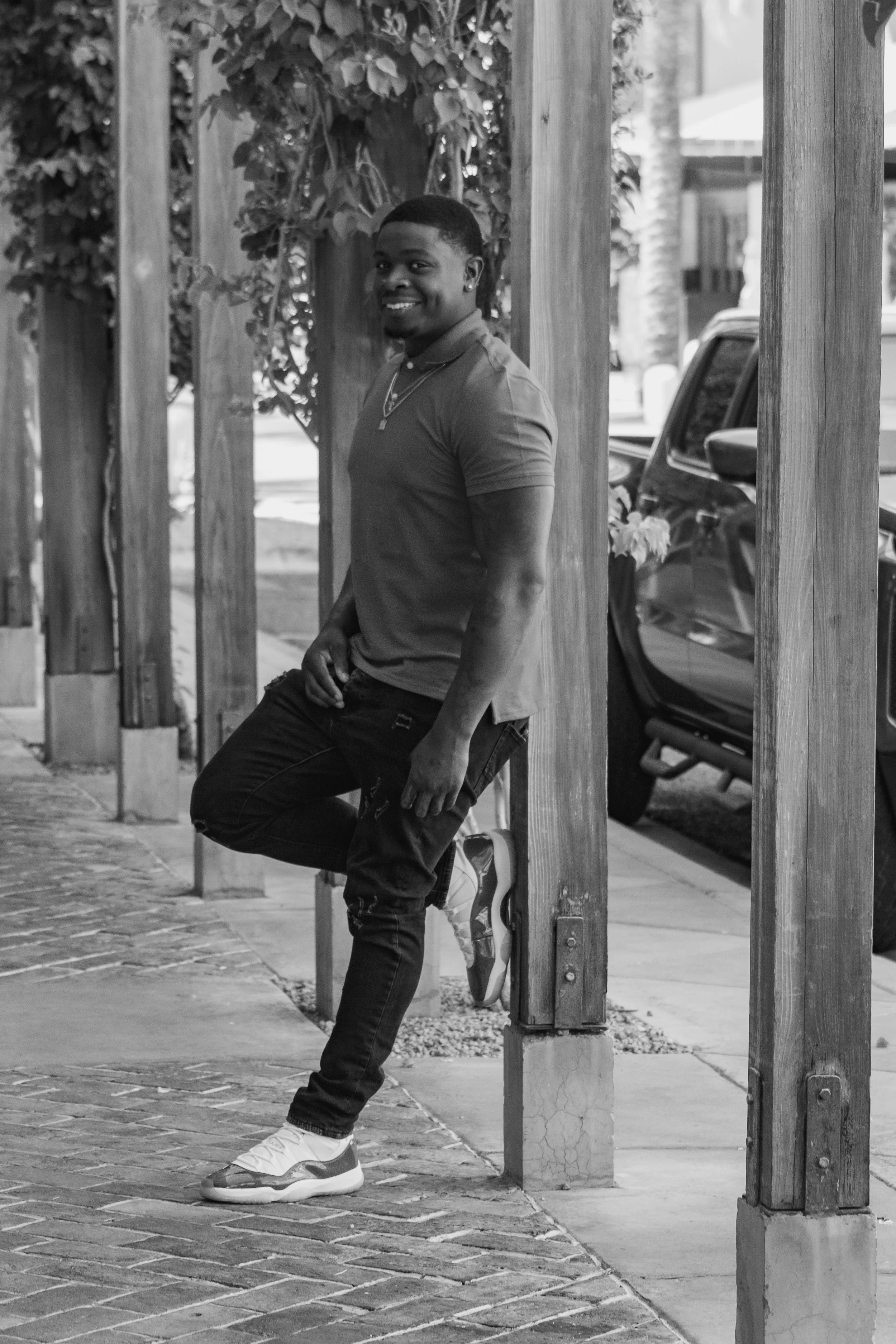 A young man with a smile, wearing a polo shirt, jeans, and sneakers, sitting and leaning against a wooden fence on a city sidewalk. There are parked cars and trees in the background.