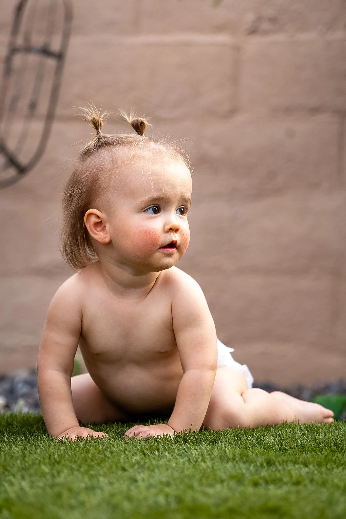 A diapered toddler with blonde hair styled in pigtails sitting on green grass in front of a brick wall, looking to the right.