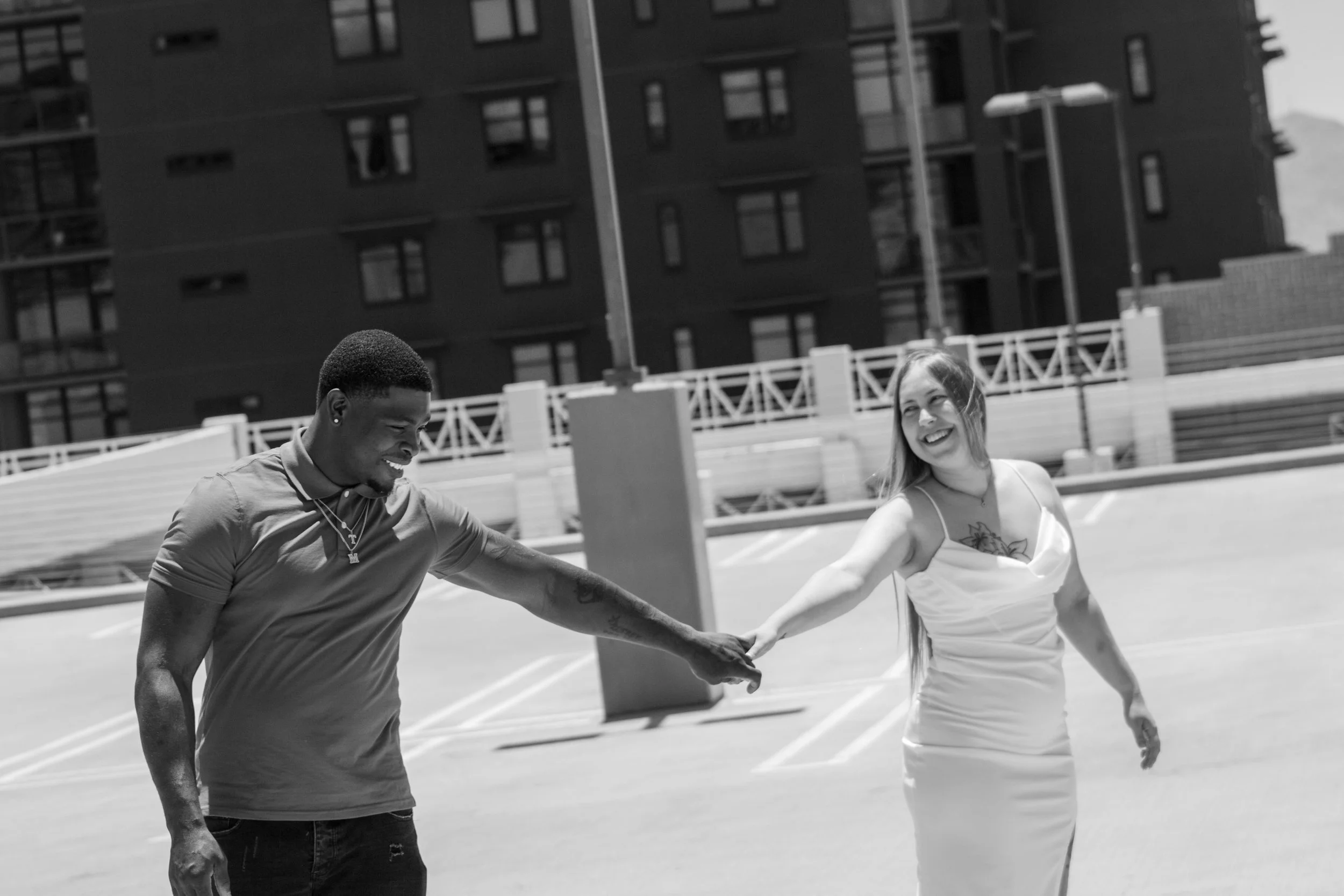 A young man and woman holding hands and smiling outdoors on a sunny day, with a building and parking lot in the background.