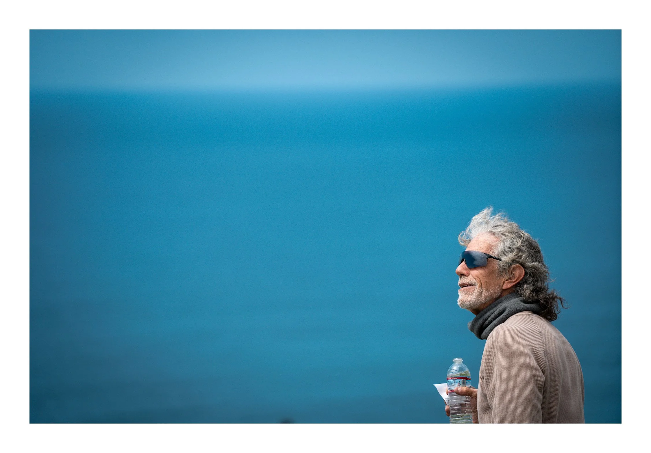 An elderly man with gray hair, wearing sunglasses, a beige blazer, and a gray scarf, standing outdoors against a clear blue sky while holding a water bottle.