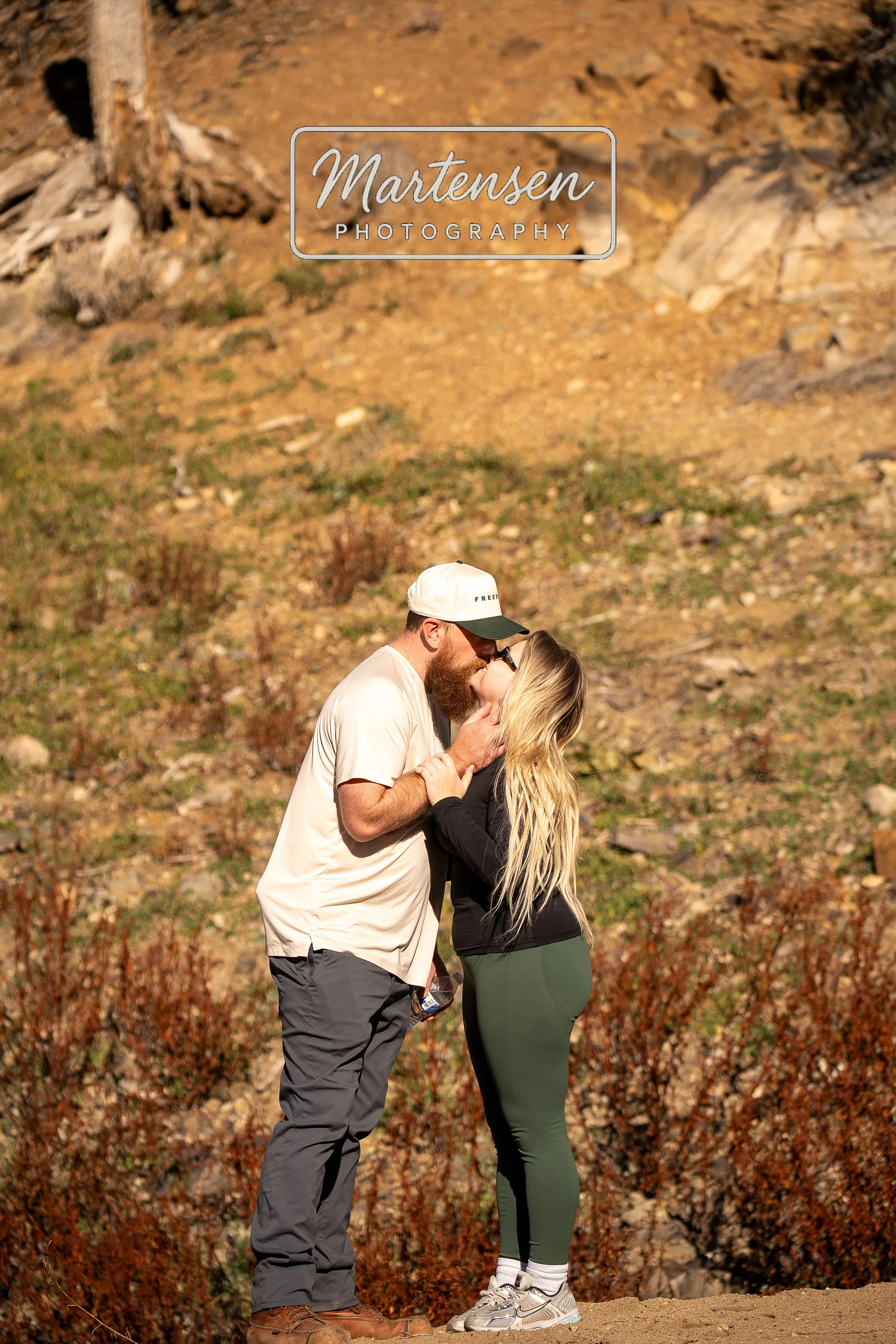 A couple stands outdoors on a dirt trail, embracing and about to kiss. The man has a beard, is wearing a white cap, a beige t-shirt, and gray pants. The woman has long blonde hair, glasses, a black top, and green leggings. The background shows a rock
