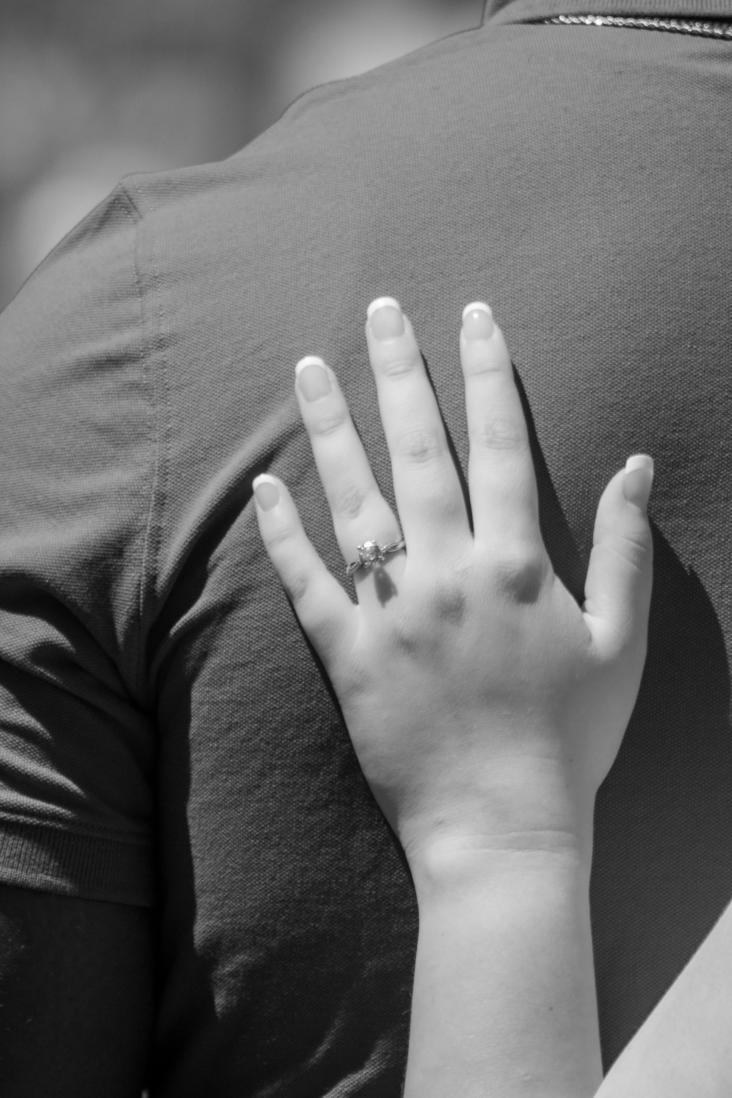 A person's hand with an engagement ring is resting on the back of someone's shoulder, both in black and white.