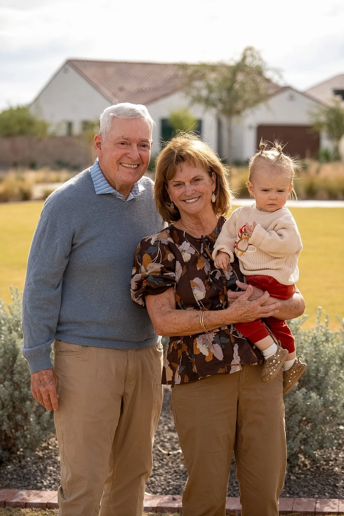An elderly man and woman smiling and standing outdoors with a young girl. The woman is holding the girl, who has light hair in pigtails and is wearing a beige sweater and red pants. The man wears a blue sweater and khaki pants. They are in a yard wit