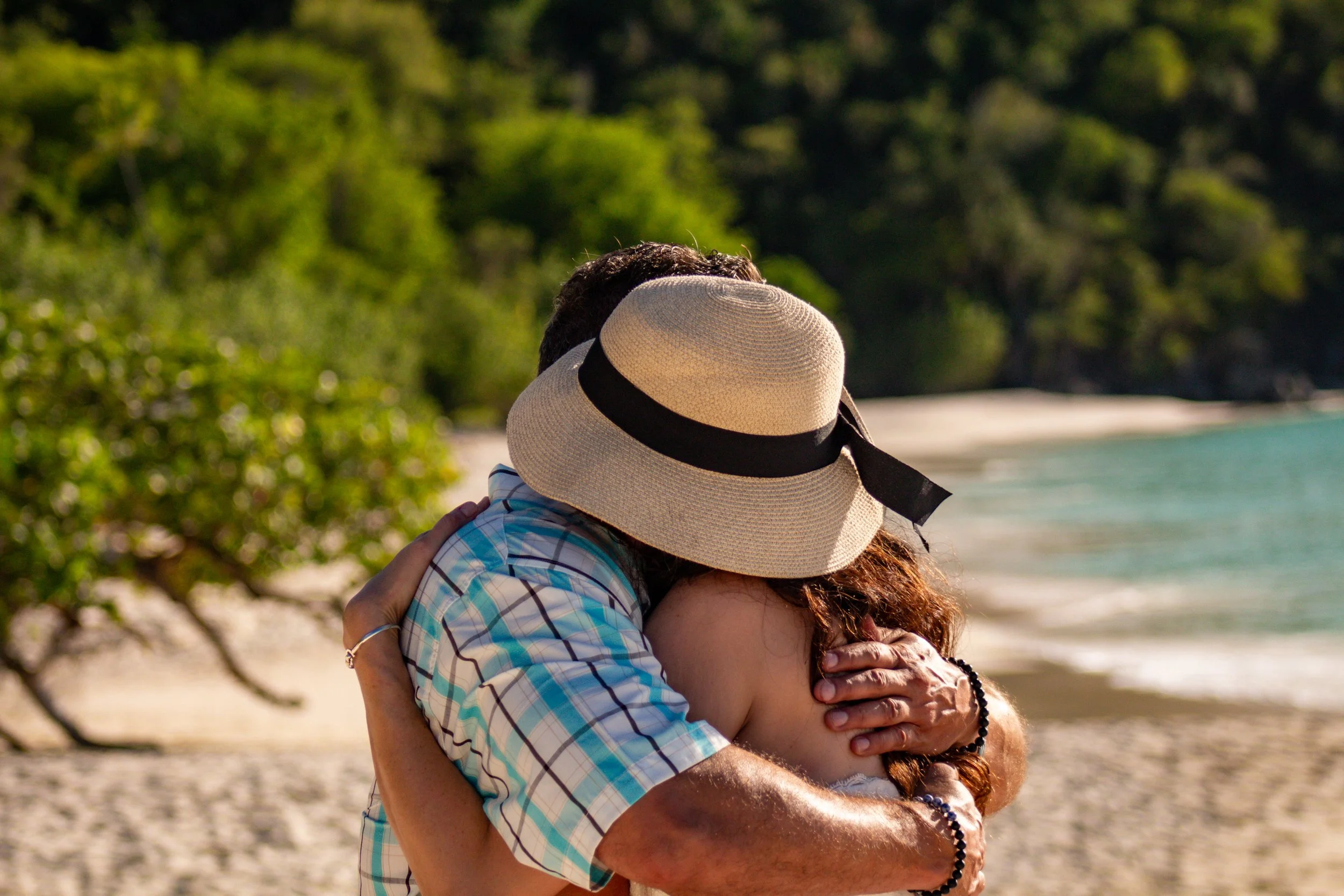 A couple hugging on a beach with trees and water in the background, one wearing a hat.