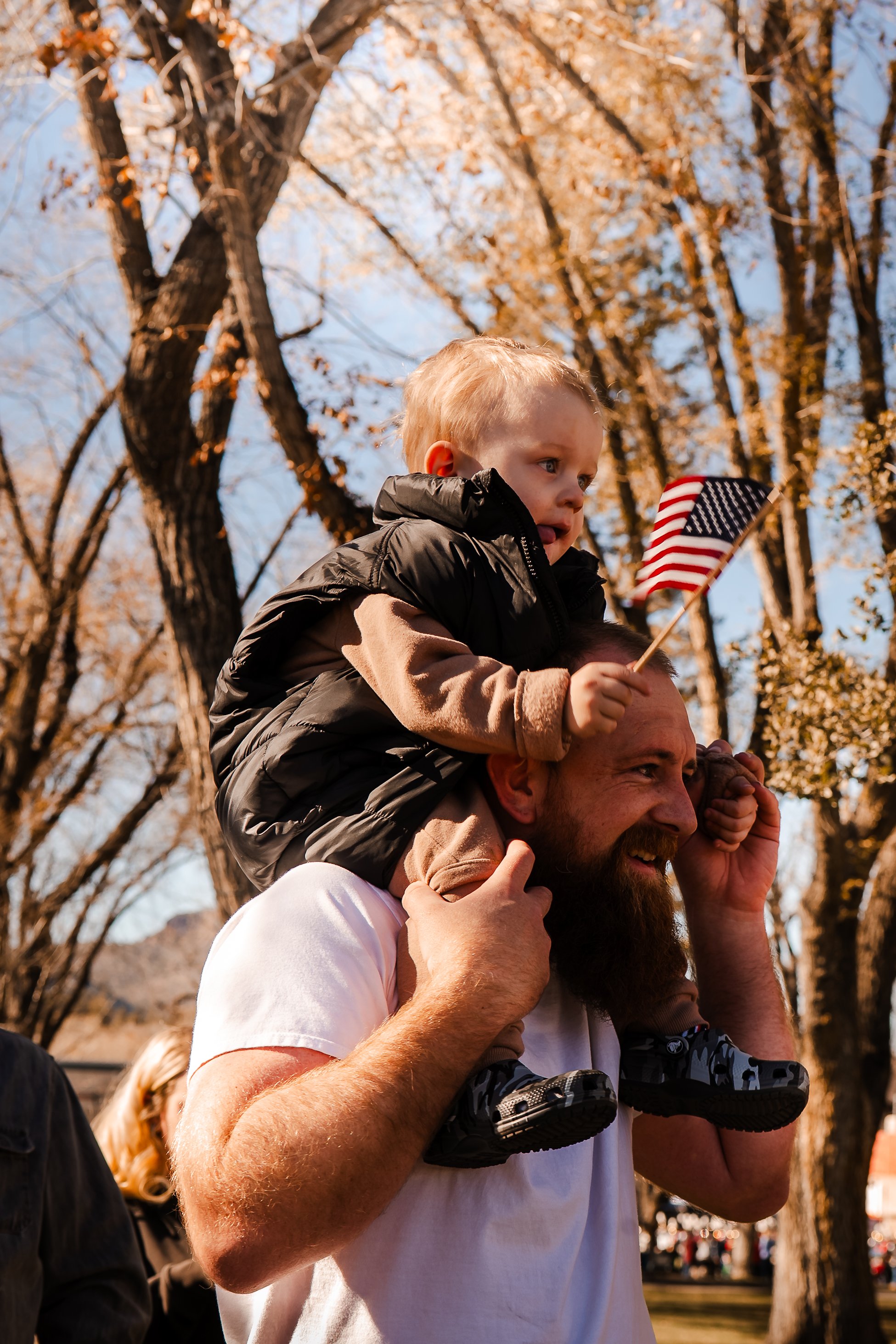 A man with a beard carrying a young boy on his shoulders outdoors during autumn, with trees and fall foliage in the background. The boy is holding a small American flag.