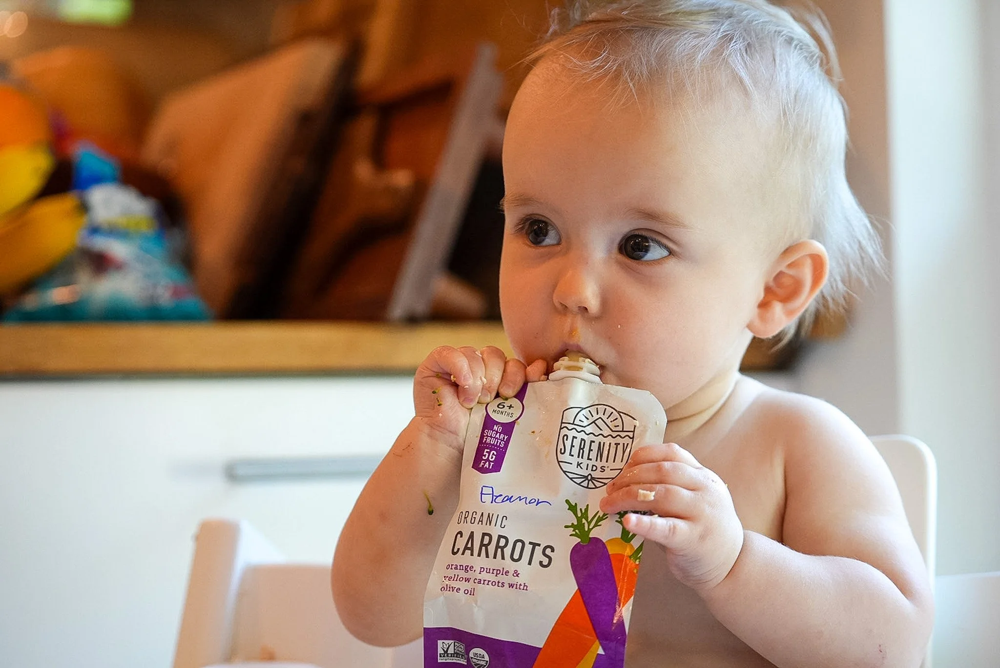 Young child with blonde hair and large blue eyes eating a pack of organic baby carrots. The child is indoors with a white high chair in front. The carrot package has colorful illustrations and text indicating it contains organic carrots and is suitab
