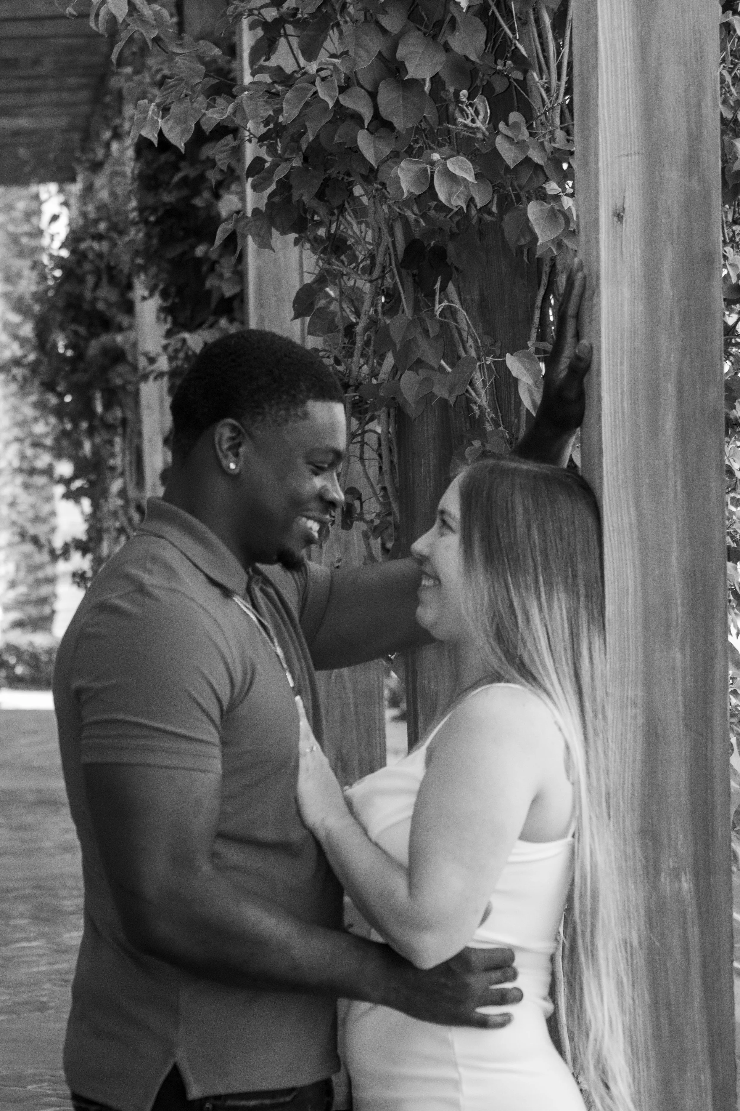 A black and white photo of a man and woman standing close to each other outdoors, with the man having his arm around the woman and their foreheads touching, smiling at each other. They are near a wooden post and leafy vines.