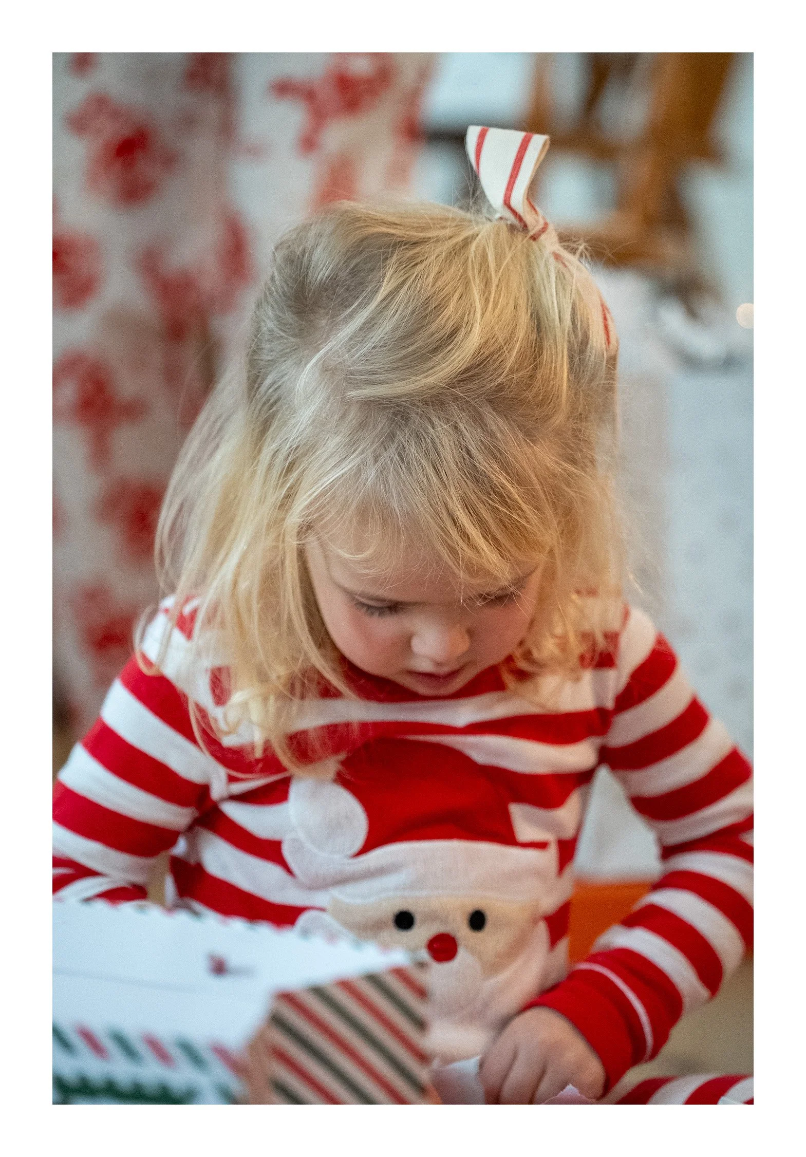 A young girl with blonde hair wearing a red and white striped Christmas sweater with a snowman face on it, opening a holiday gift.