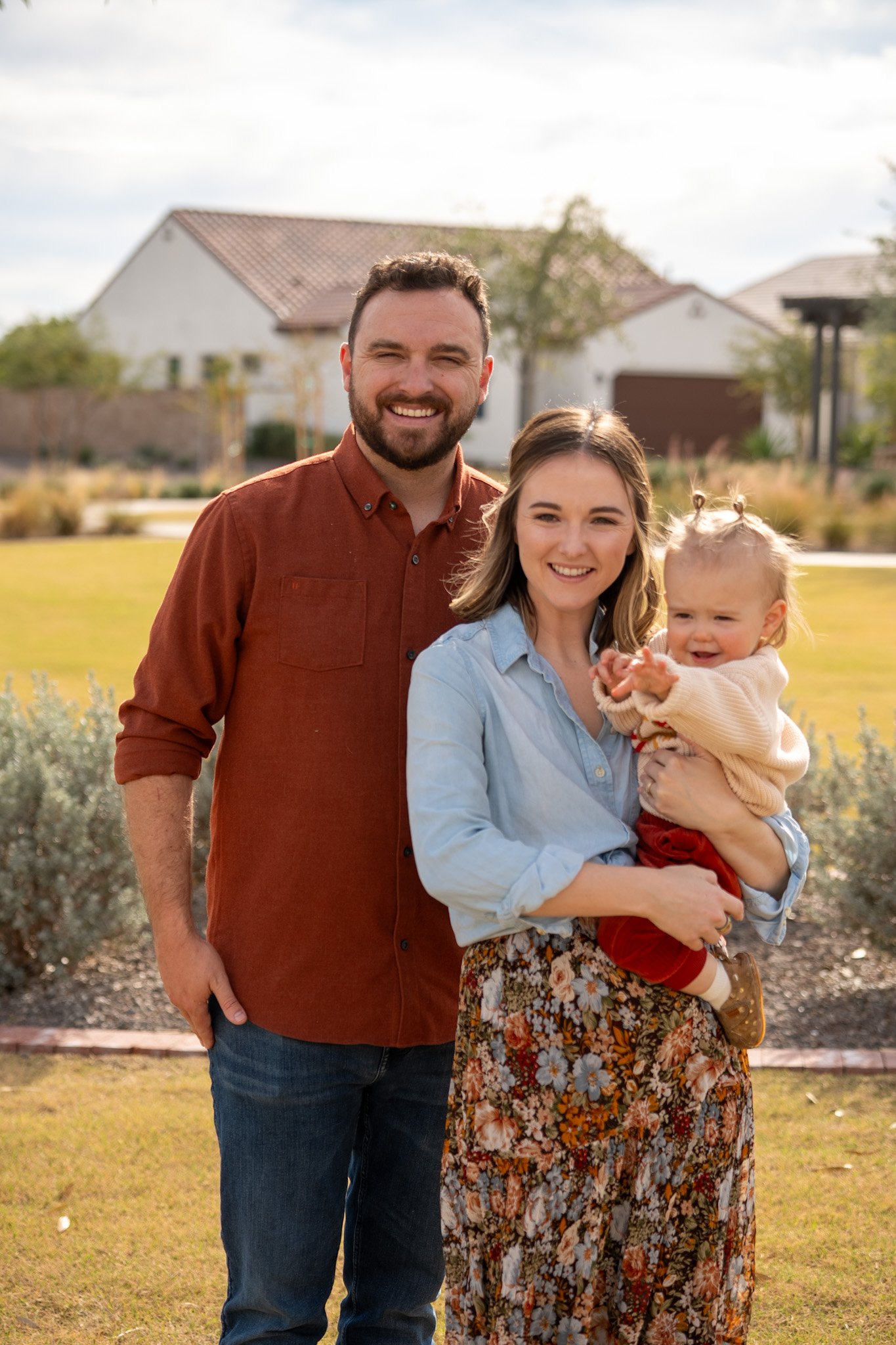 Family standing outdoors on a sunny day in front of houses and greenery, smiling at the camera.