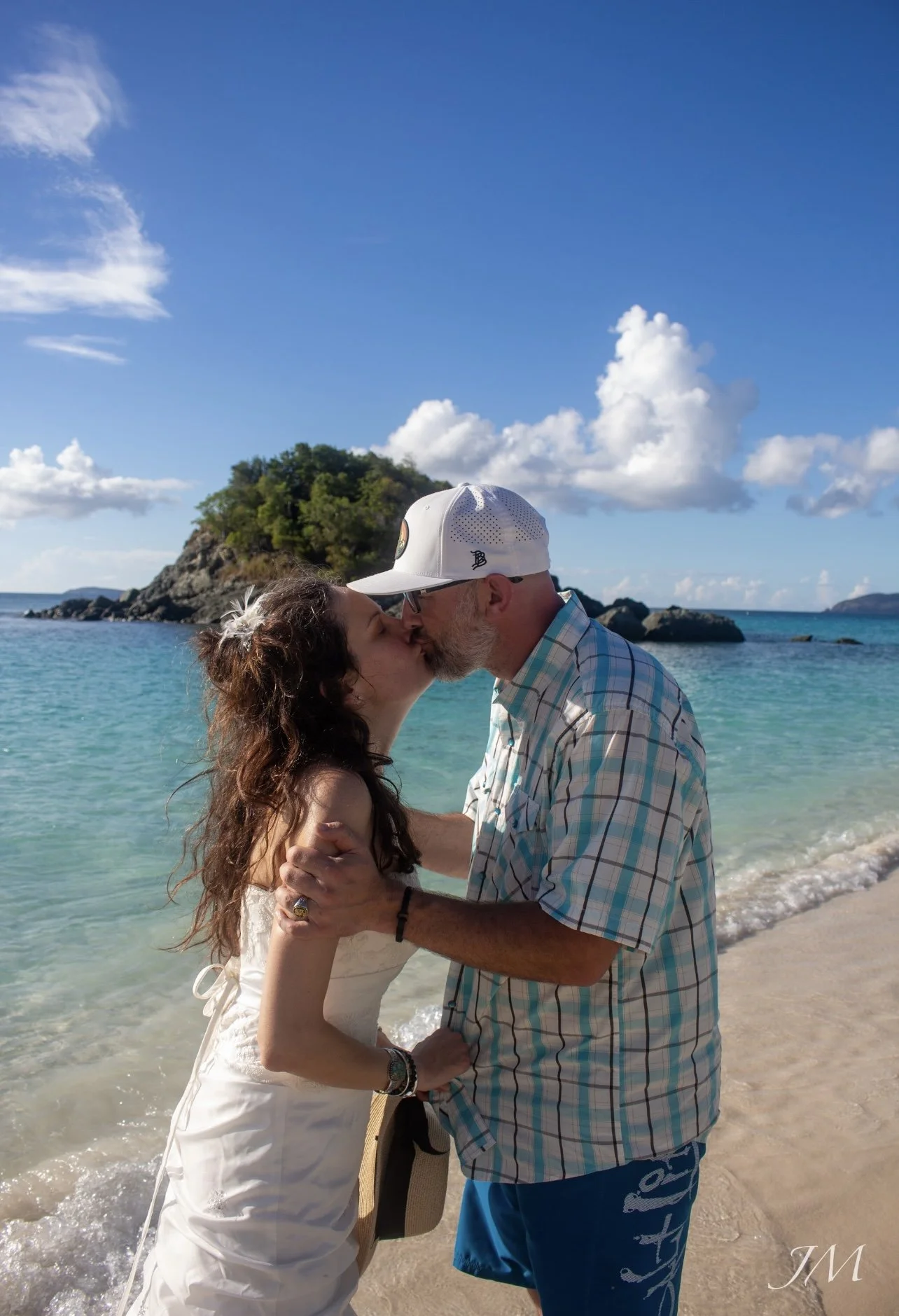 A couple kissing on the beach with ocean water and a small island behind them, under a partly cloudy sky.