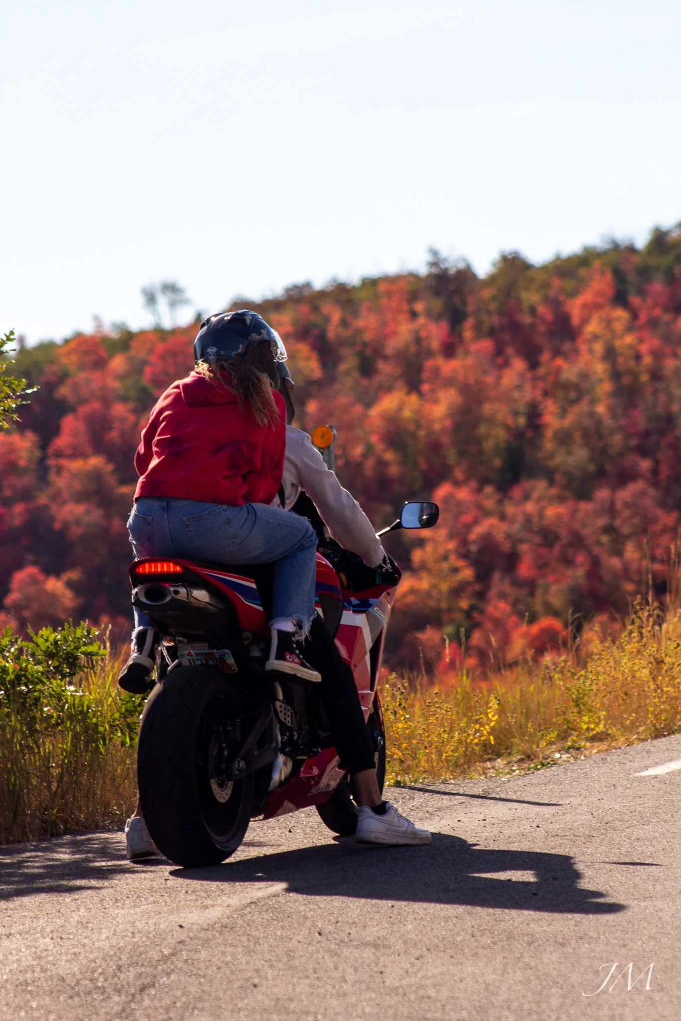 Person wearing a helmet and red hoodie riding a red and white motorcycle on a road with autumn-colored trees in the background.