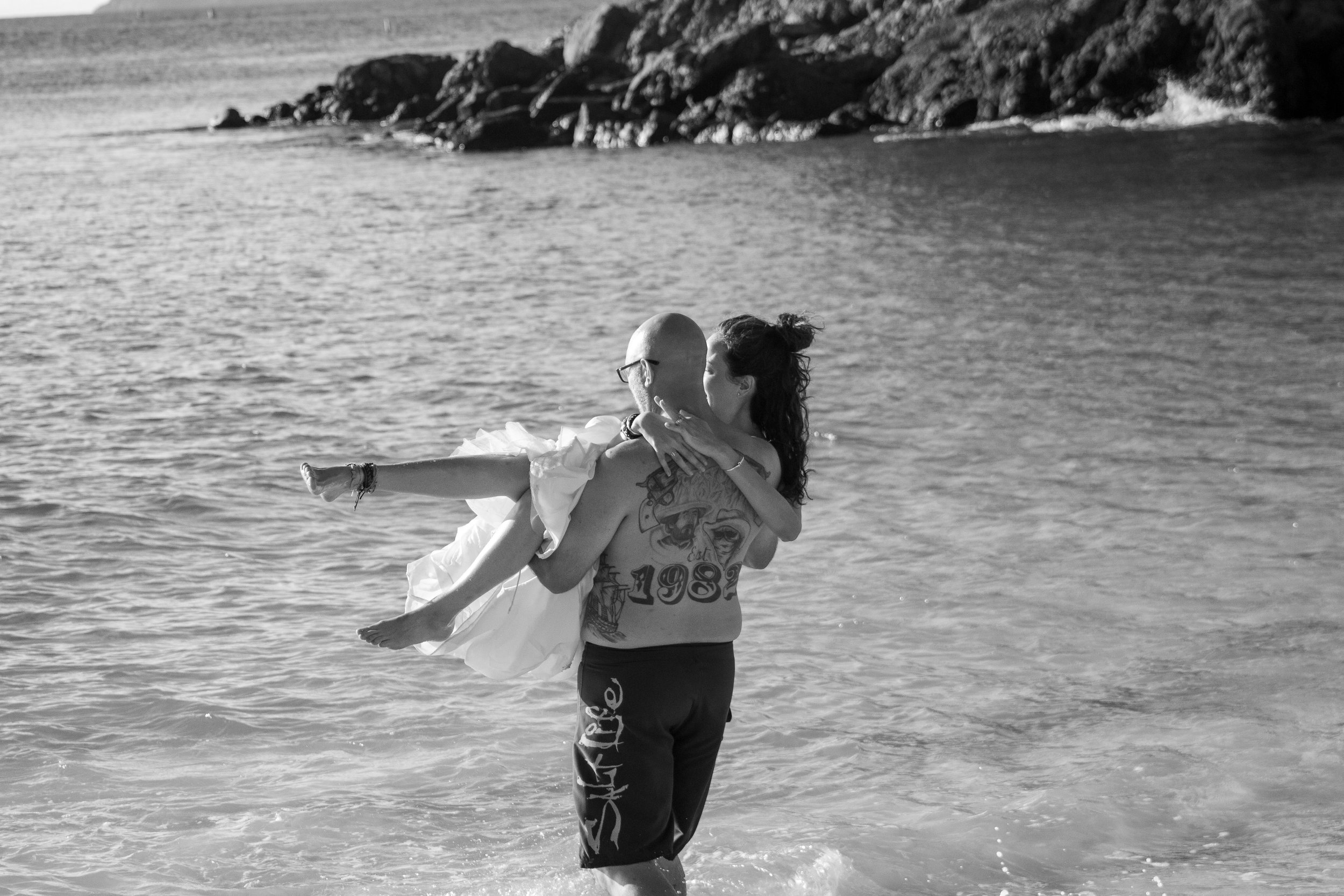 A man with tattoos carrying a woman in a dress in the ocean near rocks.
