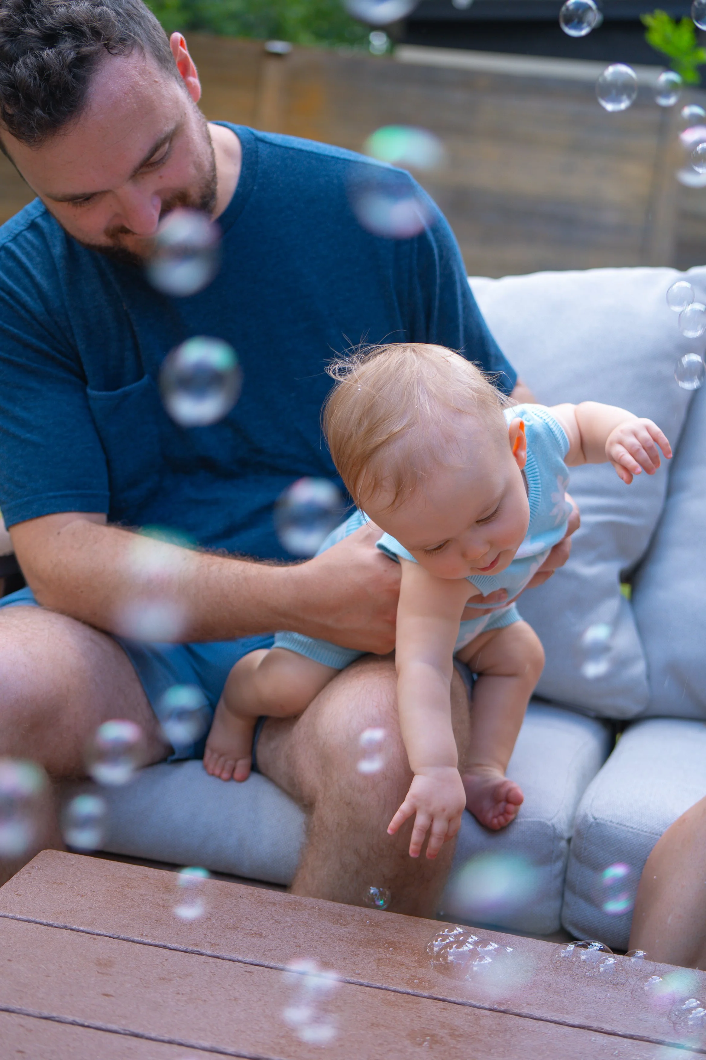 A man holding a baby outdoors on a couch, with soap bubbles floating around them.