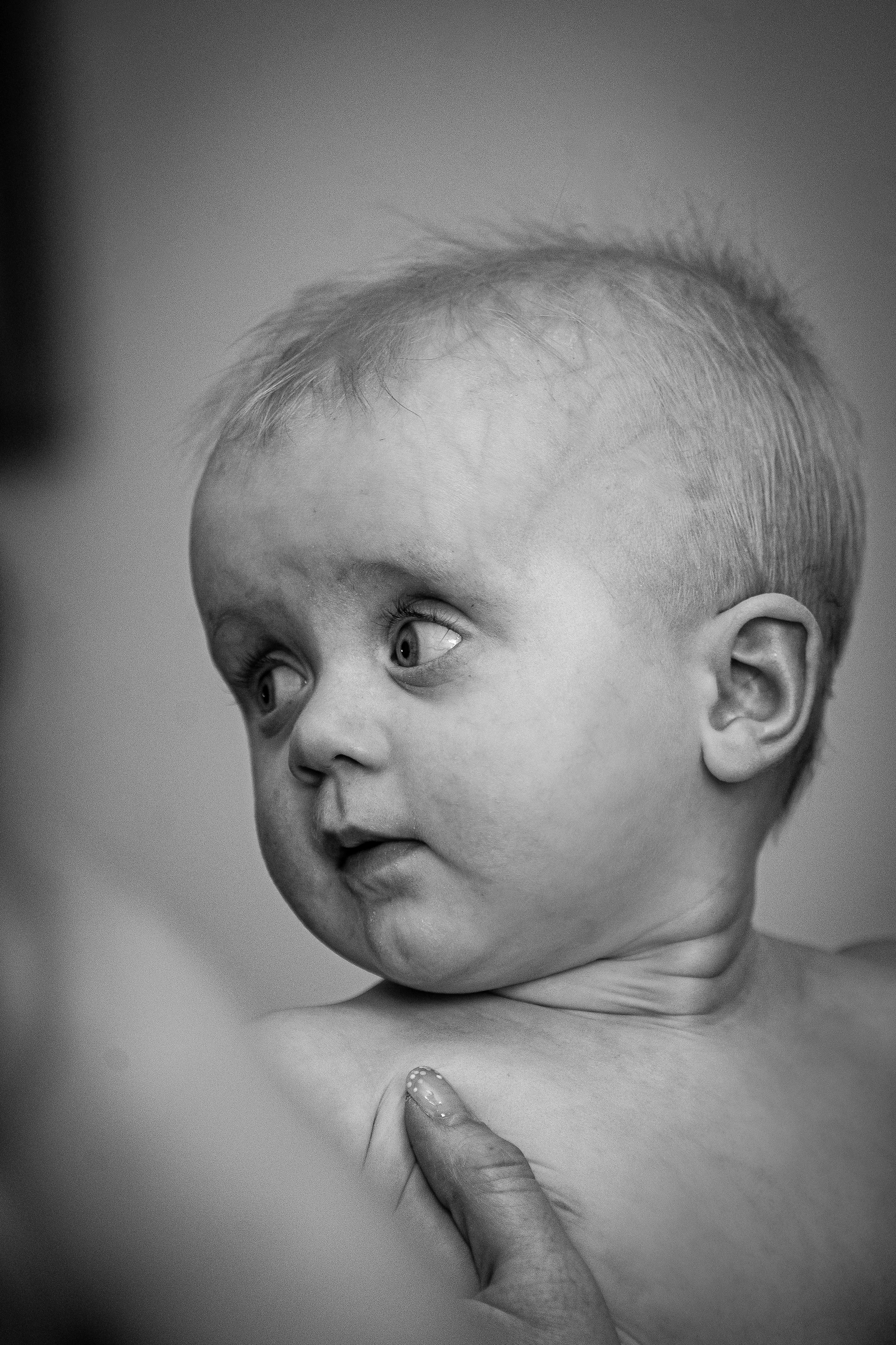 Black and white close-up of a young child's face with an expression of curiosity, looking to the side. An adult's hand is gently holding the child's chest near the shoulder.