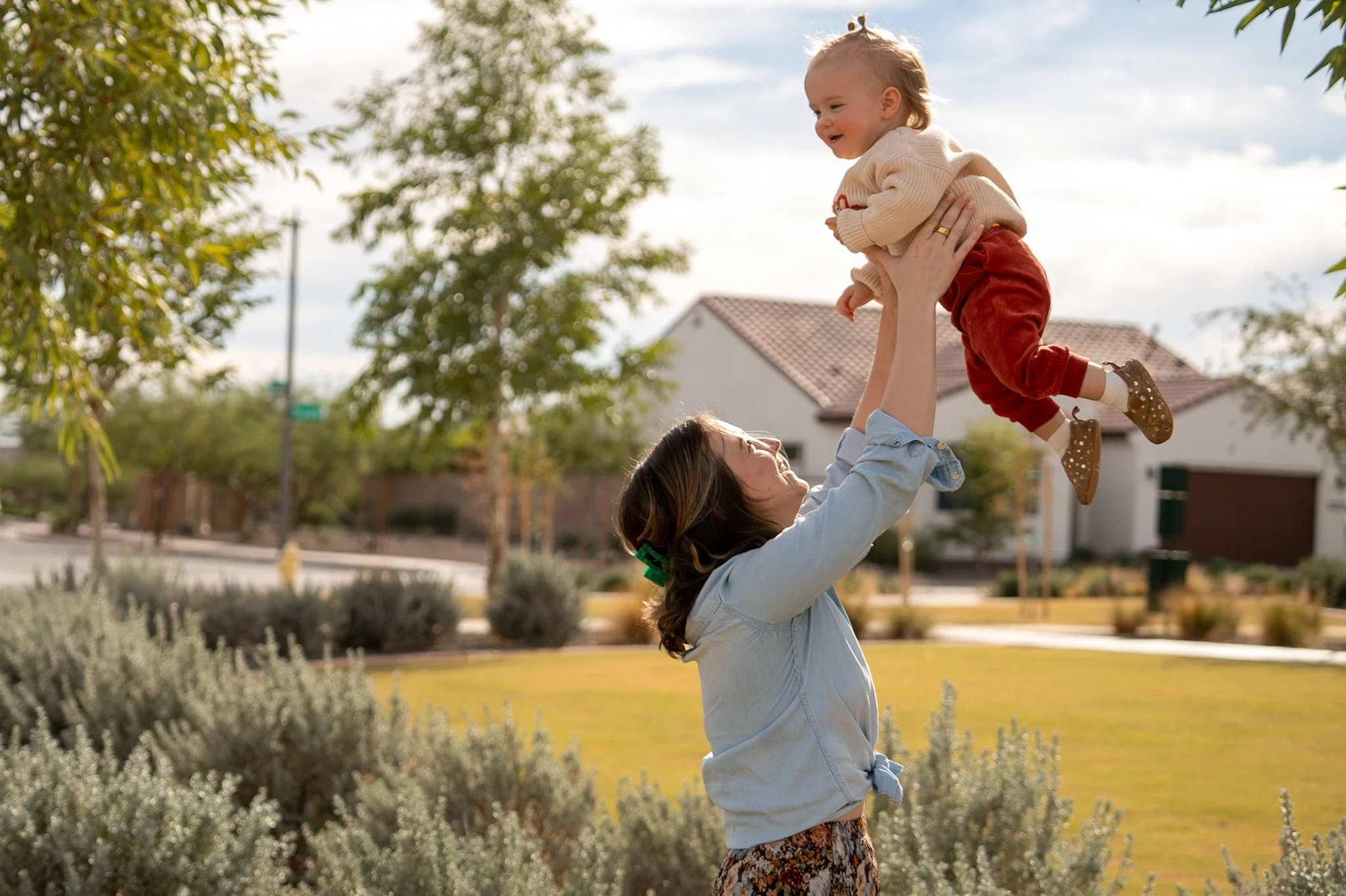 A woman lifting a young girl in the air outdoors in a park-like setting with trees and houses in the background, the girl smiling and wearing a beige sweater and red pants.