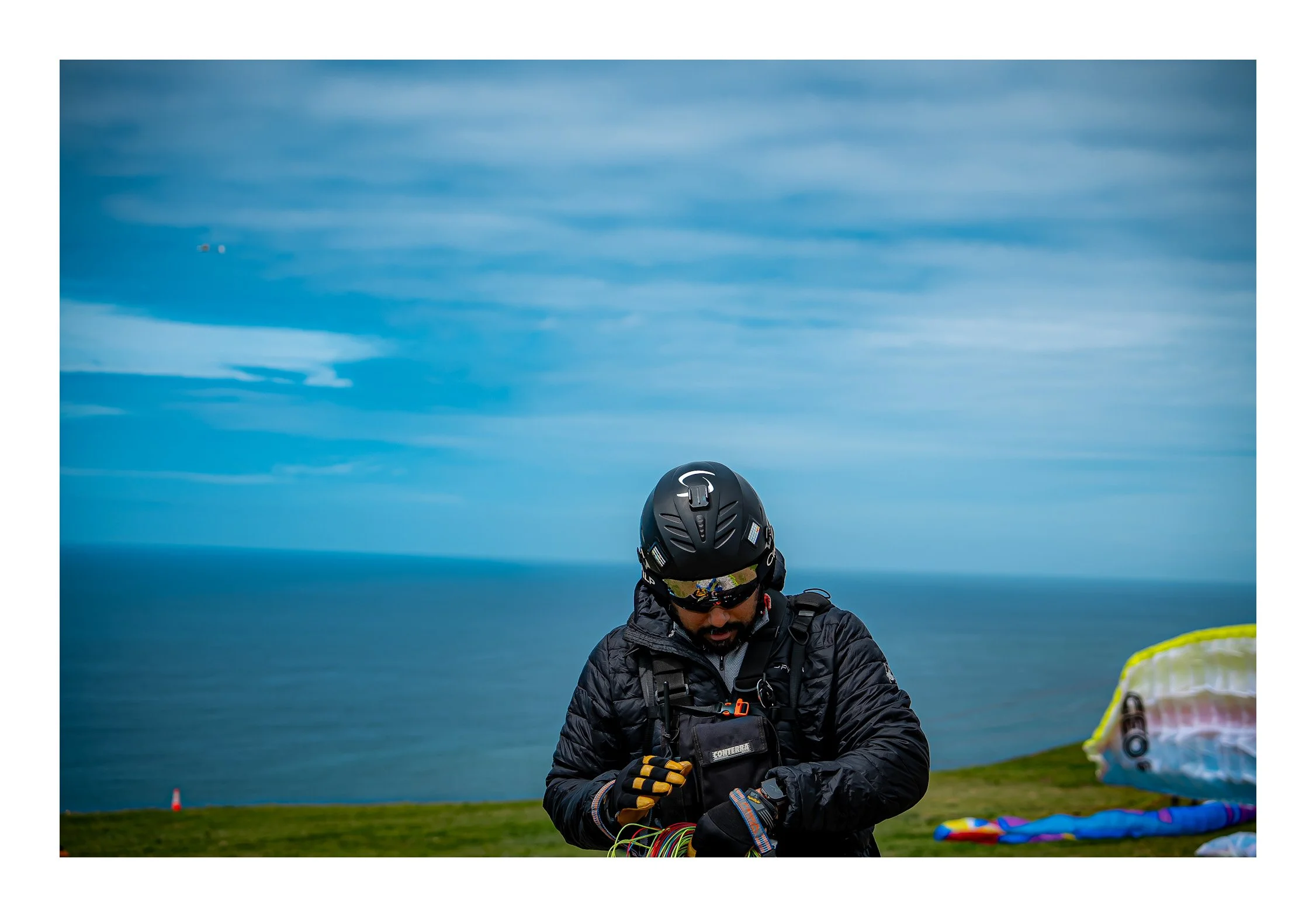 A person wearing a helmet and sunglasses, dressed in a black jacket, is outdoors with a cloudy sky in the background. They are handling electronic equipment, possibly preparing for an activity like kite flying or drone operation. Various colorful ite