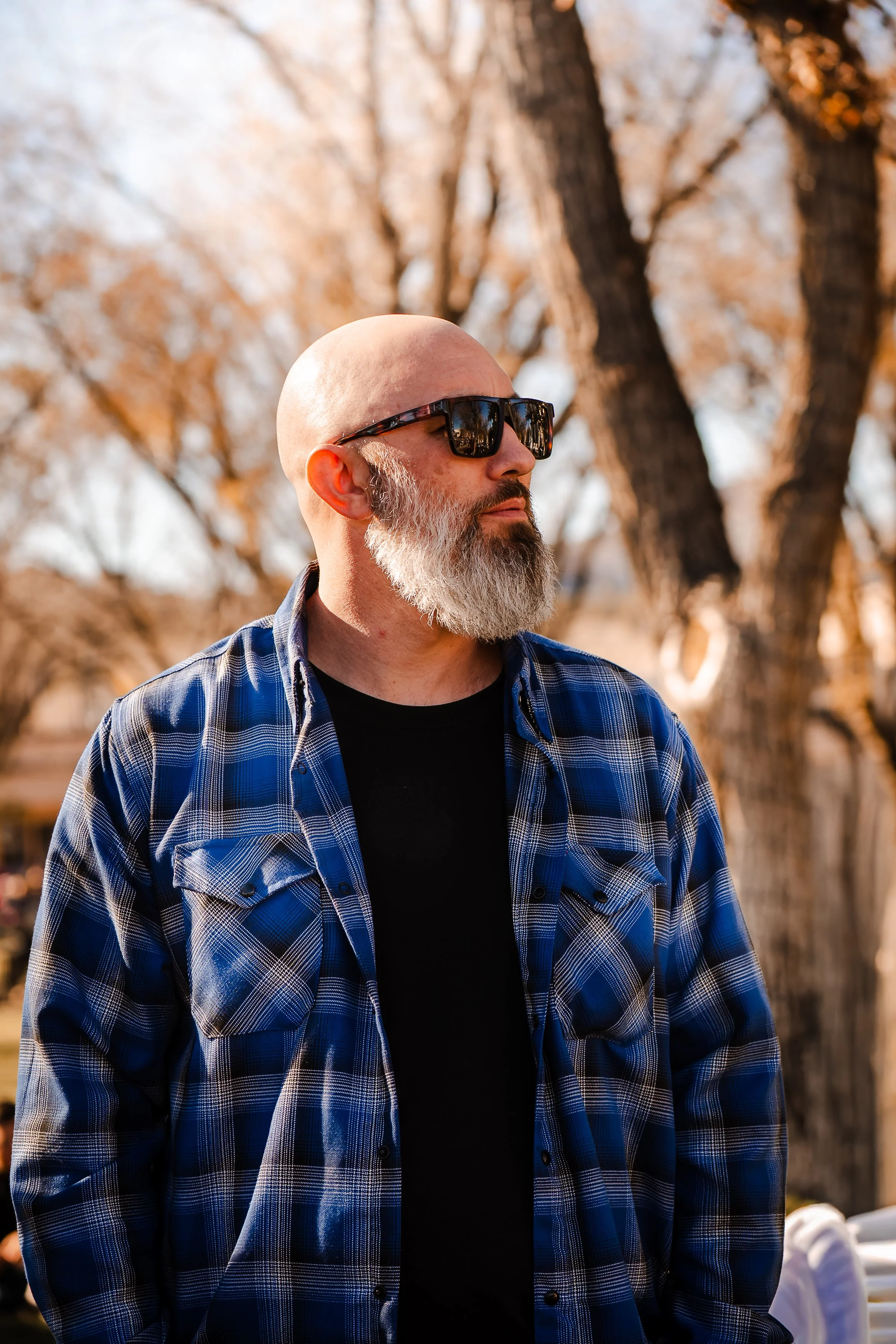 A bald man with a gray beard wearing sunglasses and a blue plaid shirt over a black T-shirt, standing outdoors with blurred autumn trees in the background.