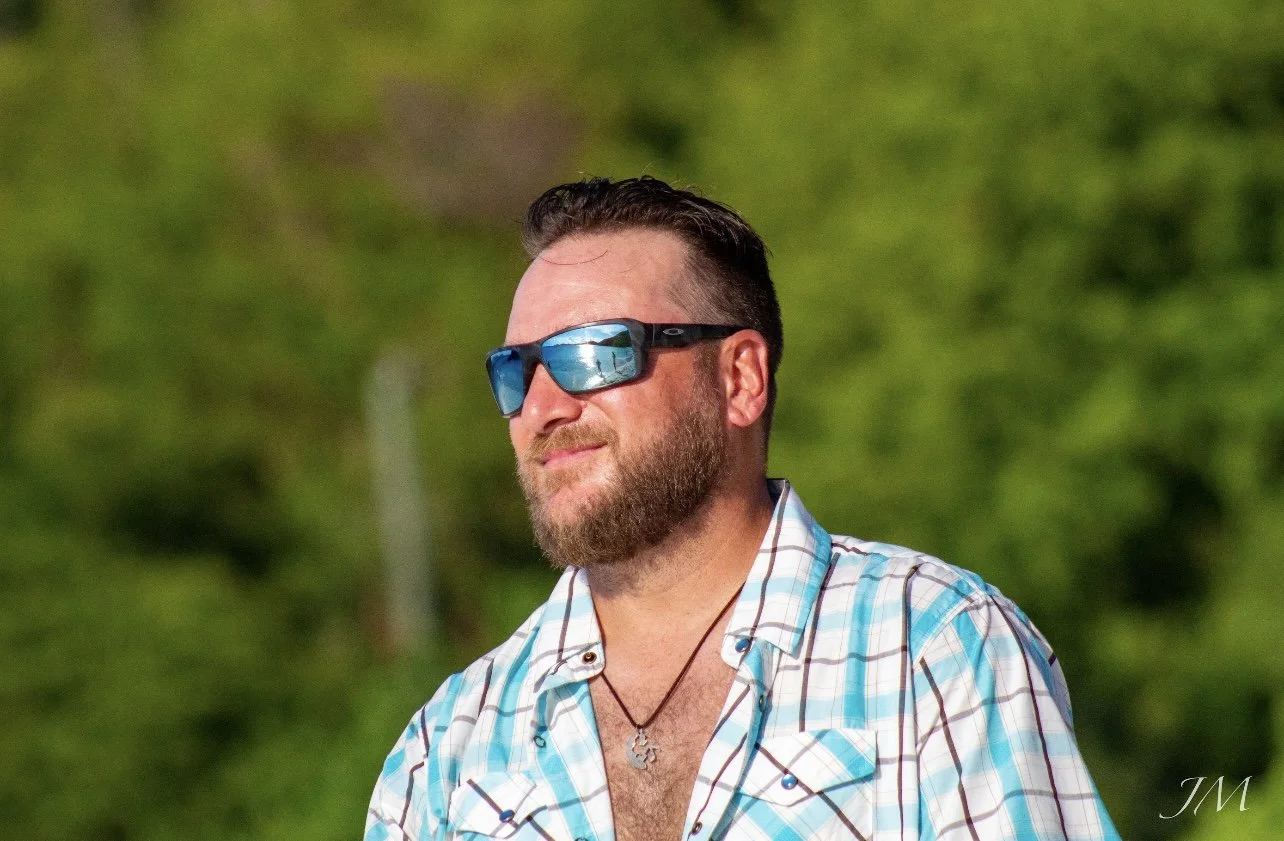 A man with a beard wearing sunglasses and a plaid shirt outdoors with green trees in the background.