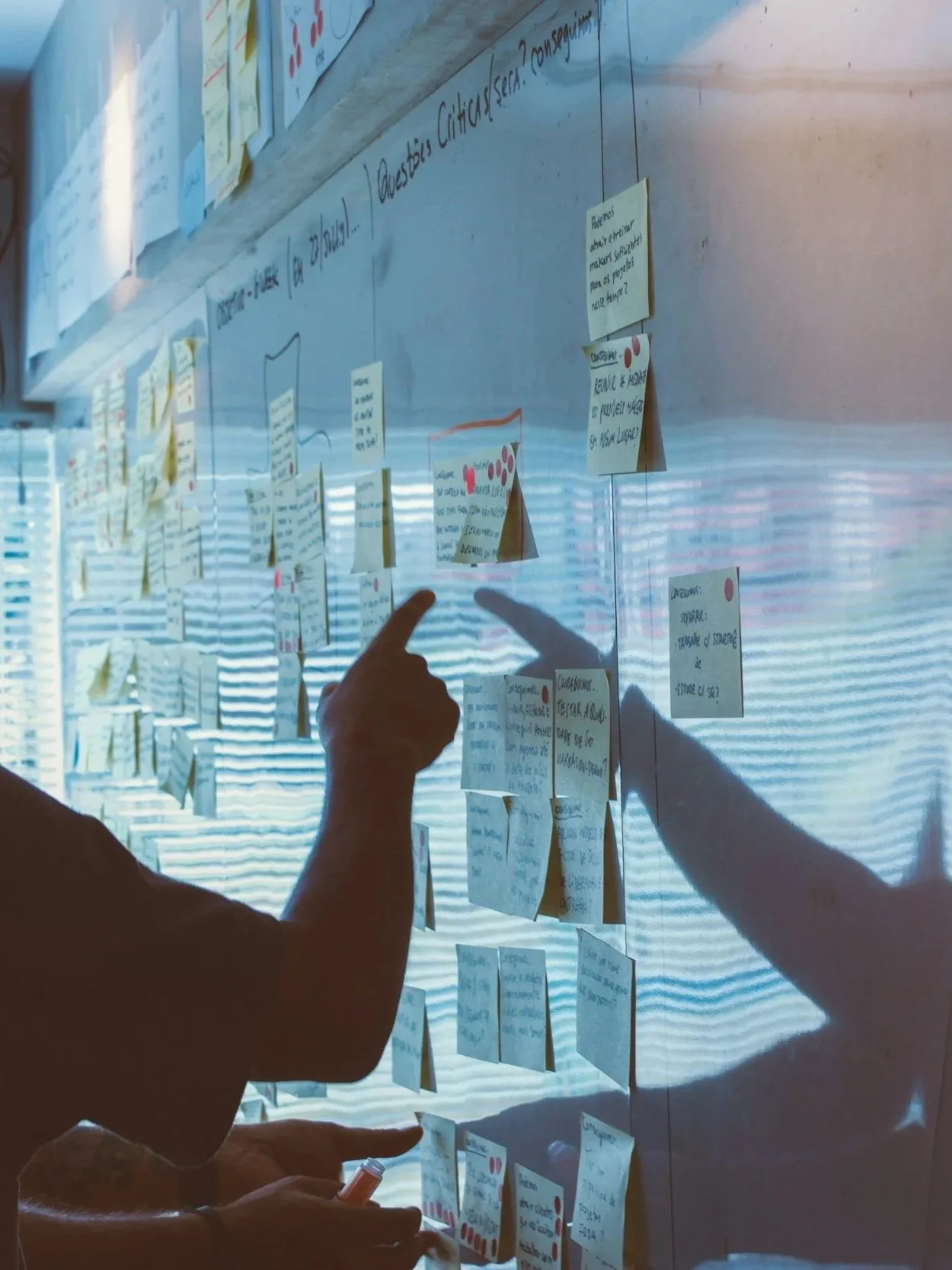 Two people are pointing at sticky notes on a glass wall during a collaborative planning or brainstorming session.