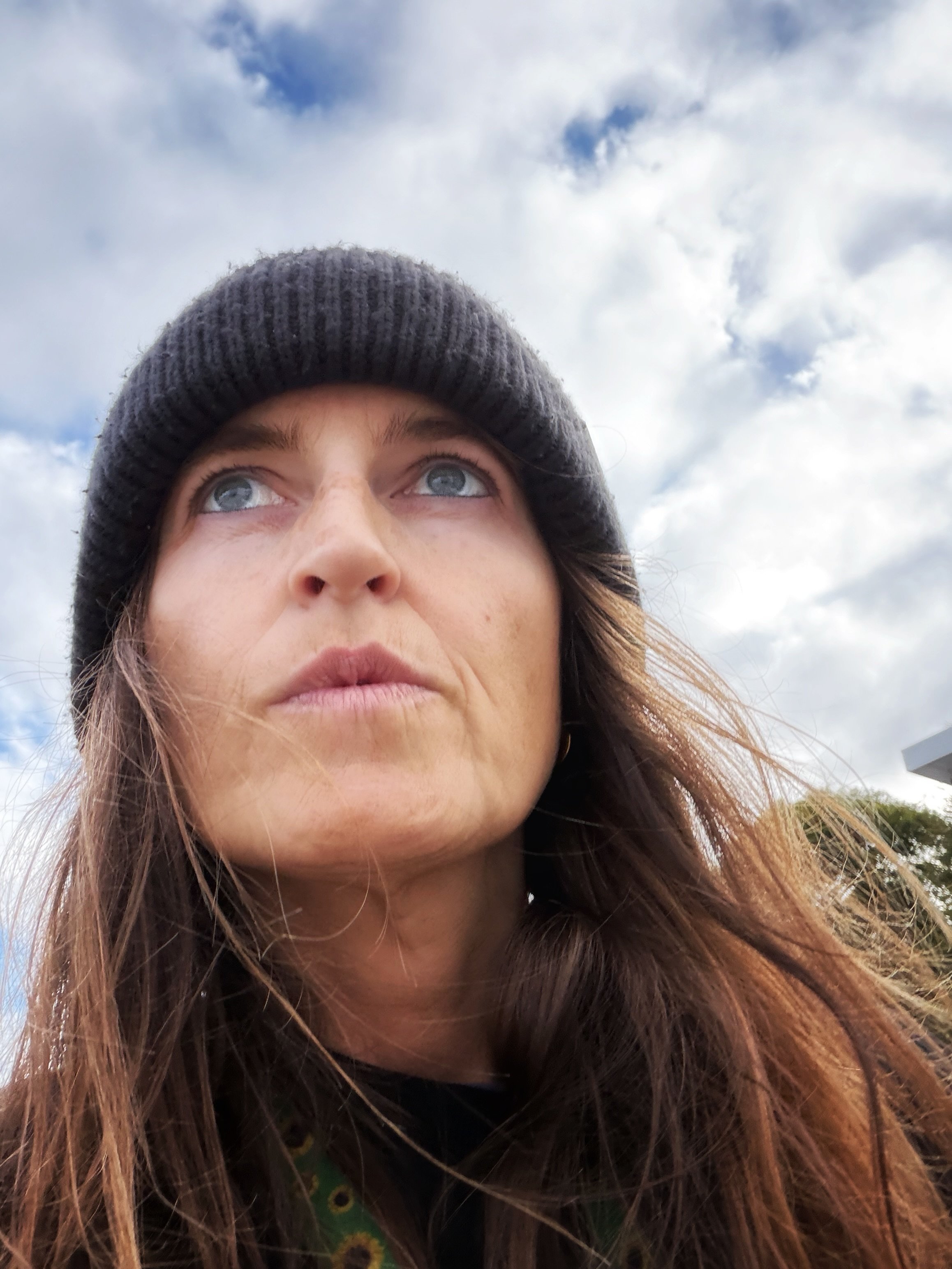 Close-up of a woman with red hair wearing a dark beanie hat, looking upward, with cloudy sky in the background.