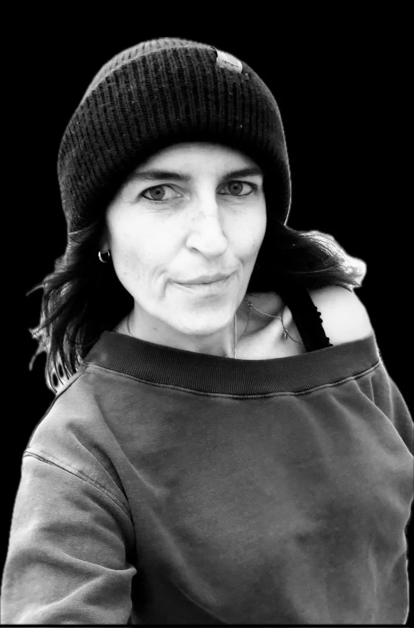 Black and white photo of a woman wearing a beanie, looking at the camera with a slight smile, against a dark background.