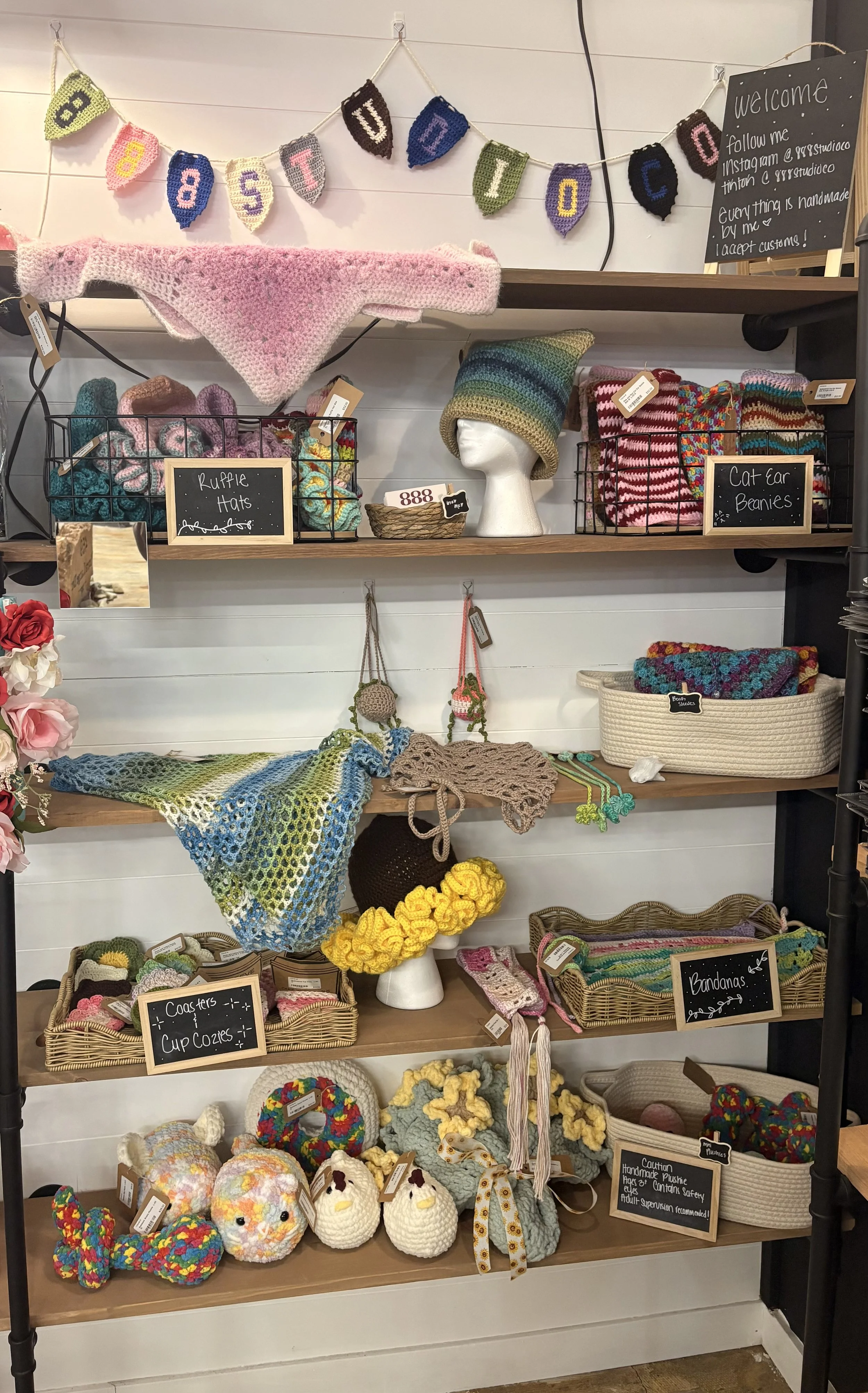 Display of various handmade knitted and crocheted items, including hats, beanies, scarves, and decorative objects, with labels and a decorative banner reading 'EAST DIO' at the top.