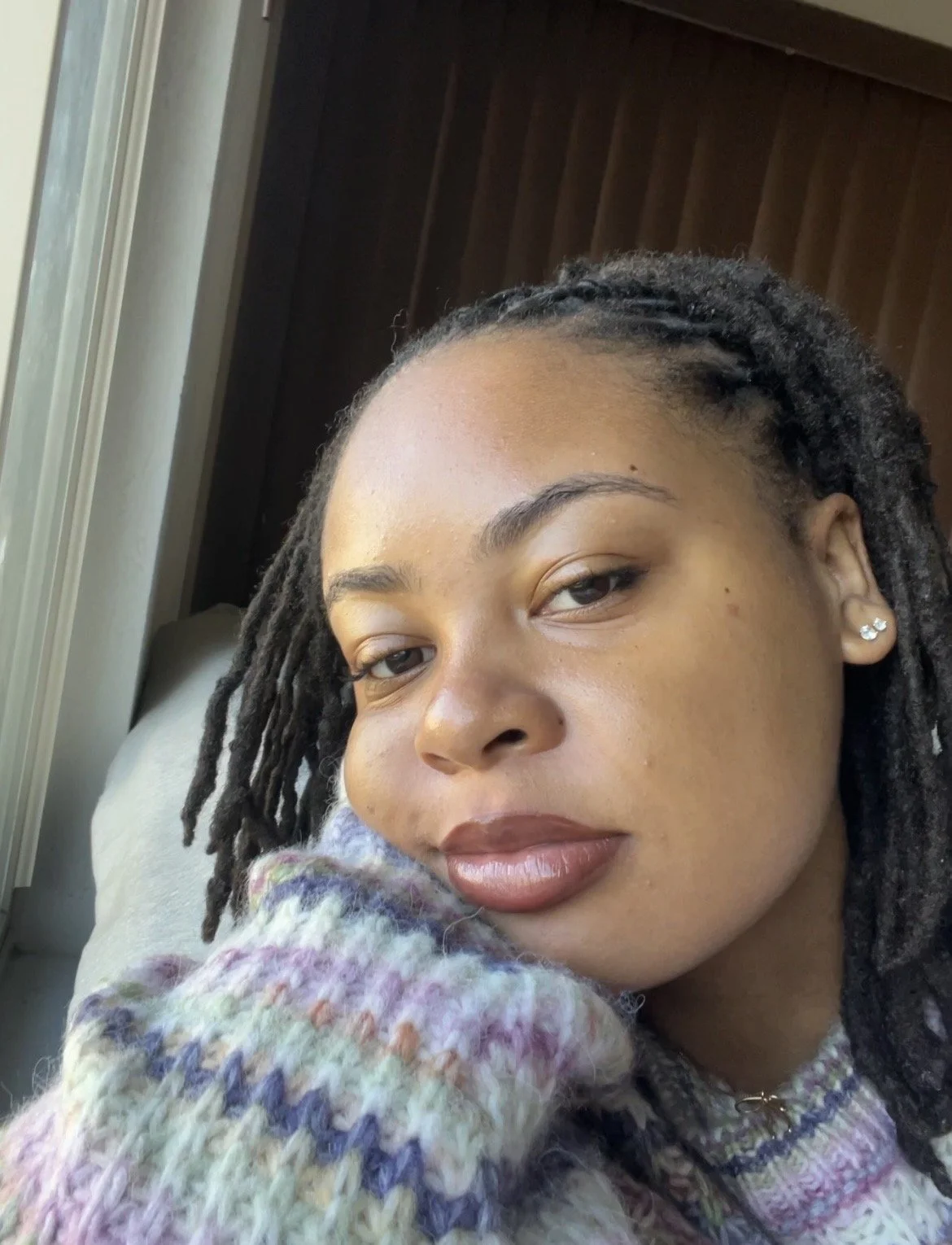 A close-up selfie of a woman with dreadlocks, wearing earrings, resting her face on her hand, standing near a window with natural light, indoors.