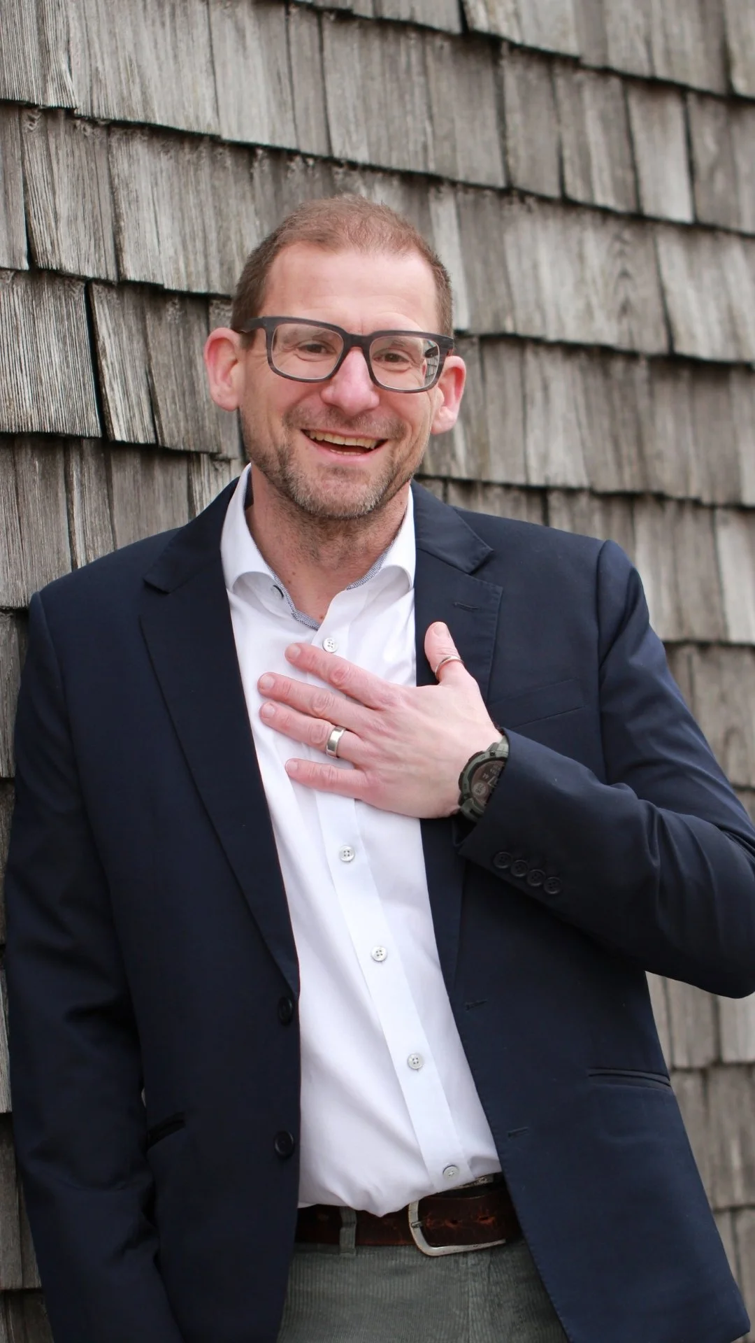 Ein lachender Mann in Brille, Anzug und weißem Hemd lehnt an einer Holzwand, die Hand auf der Brust, mit einem Lächeln.