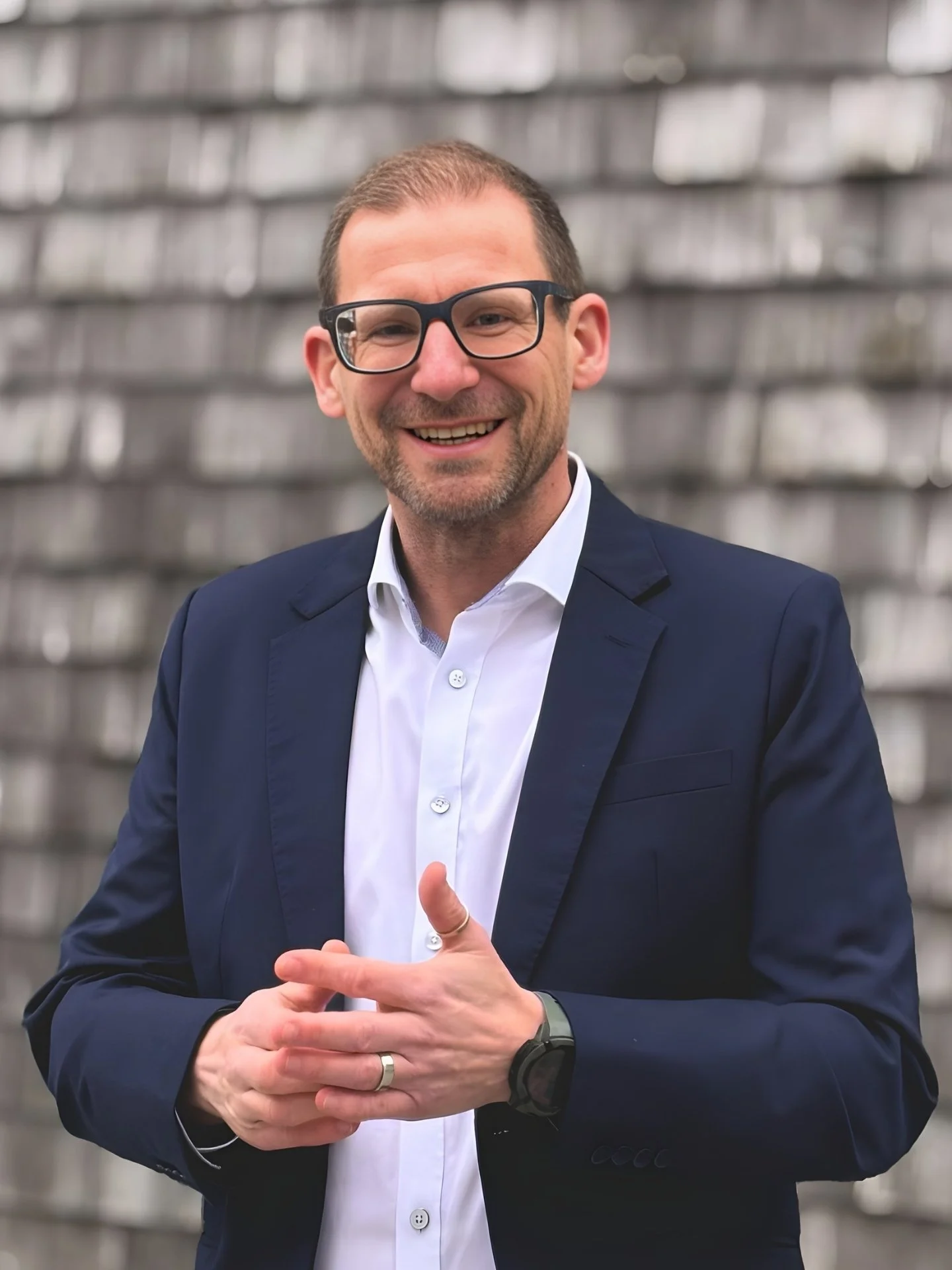 Ein lächelnder Mann in einem dunklen Anzug und weißem Hemd vor einem grauen, modernen Gebäude.