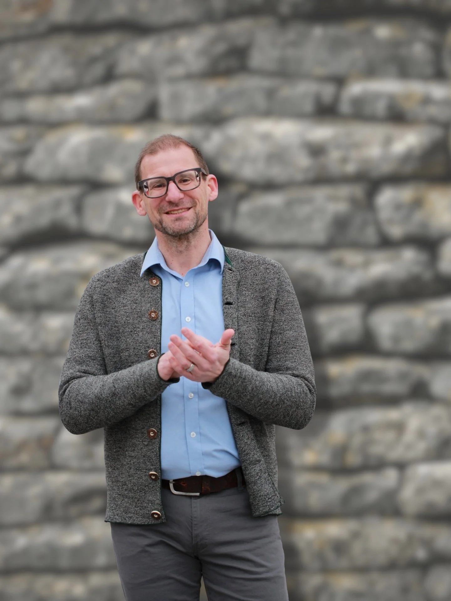Ein Mann mit Brille, grauer Jacke, blaßblauer Hemd, grauen Hosen, steht vor einer Steinmauer, lächelt und hat die Hände zusammen.