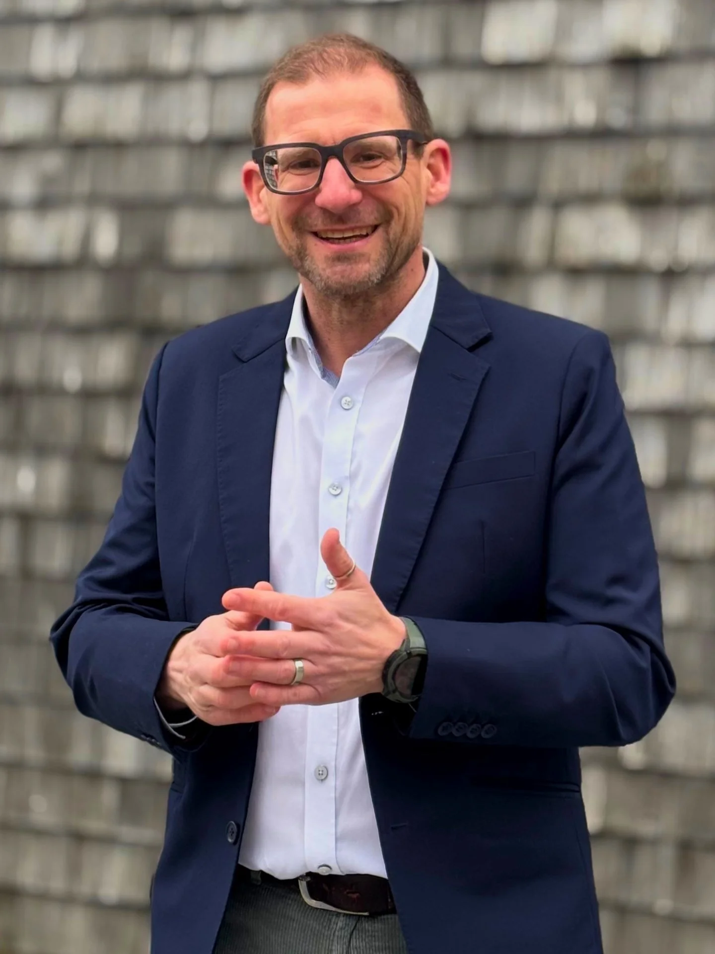 Ein lächelnder Mann in einem blauen Anzug und weißen Hemd, trägt eine Armbanduhr und eine Ehering. Er steht vor einer Ziegelwand.