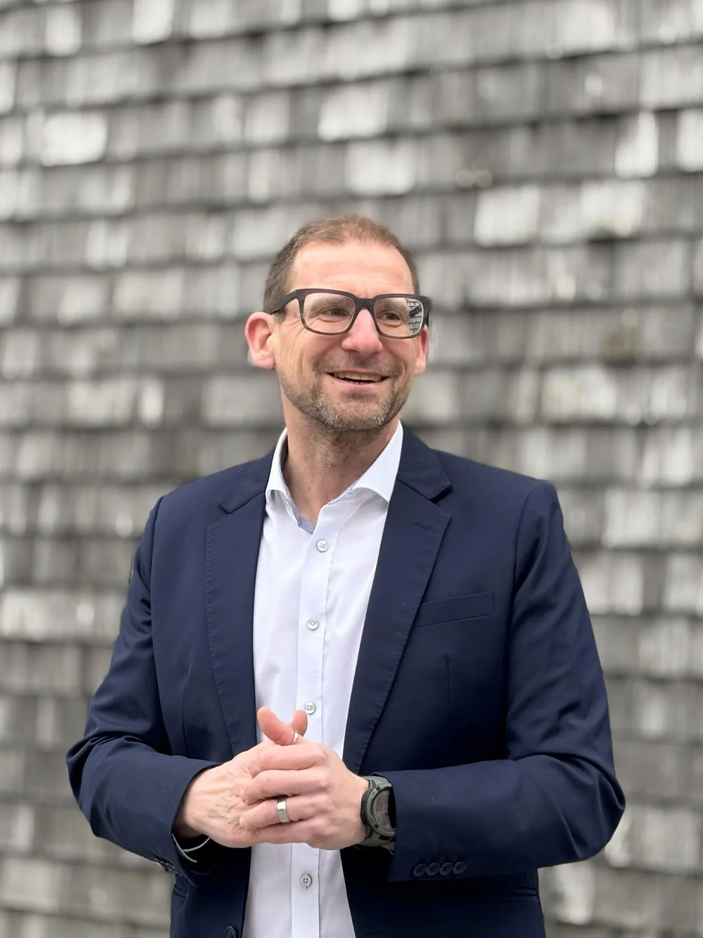 Ein lächelnder Mann in Brille, Anzug und weißem Hemd steht vor einer Wand aus Holzplanken.