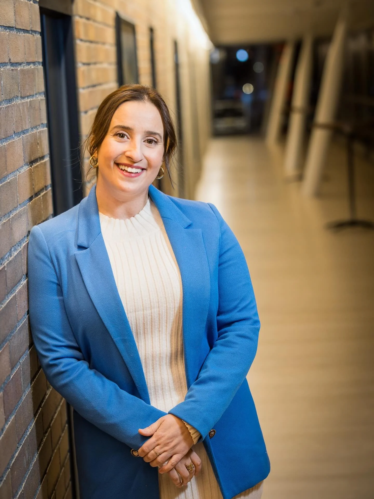 A smiling professional woman with brown hair, wearing a blue blazer and a white sweater, standing against a brick wall.
