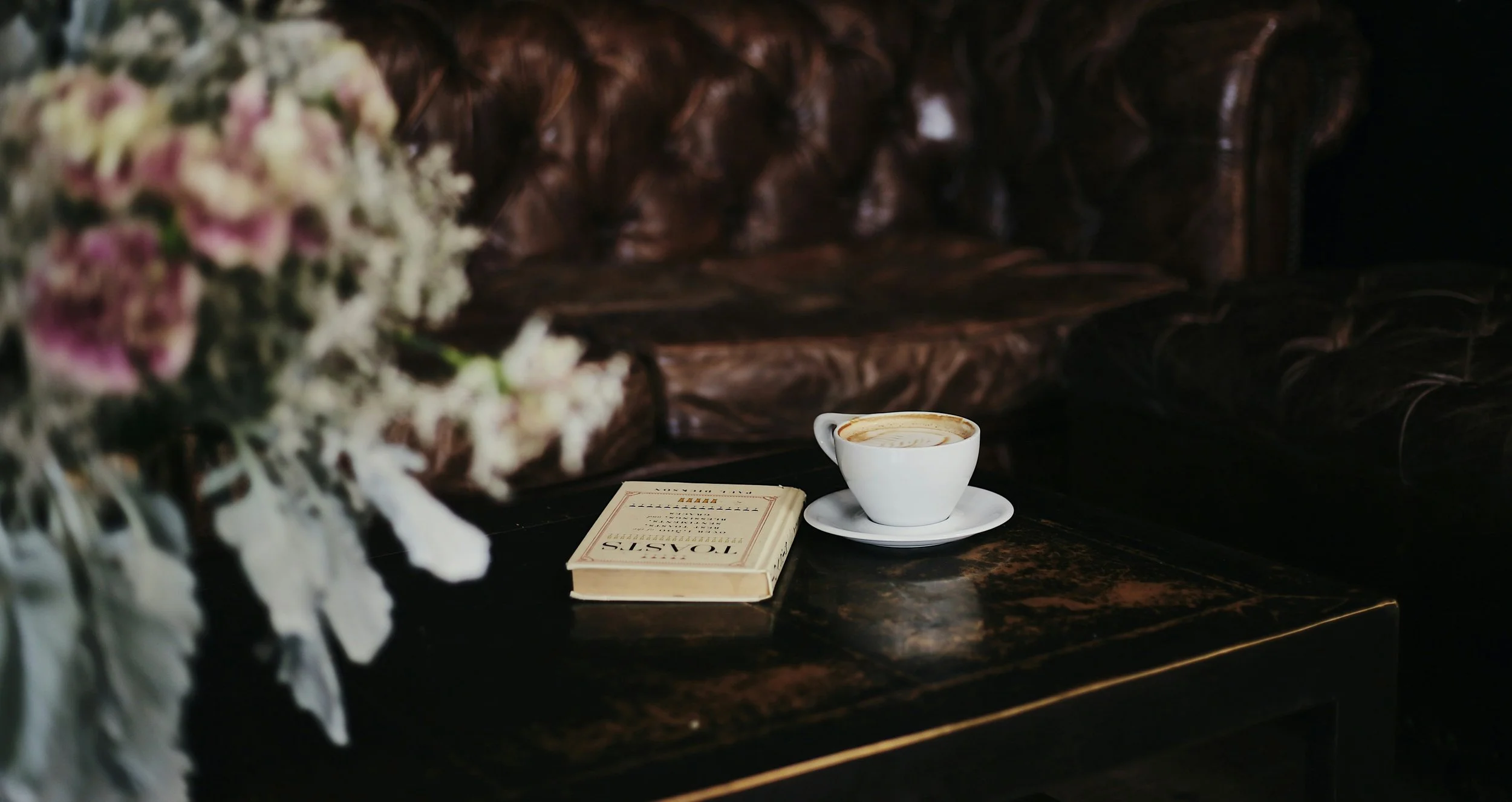 A cup of coffee on a saucer, a closed book, and a blurry bouquet of pink and white flowers on a dark, vintage leather sofa.