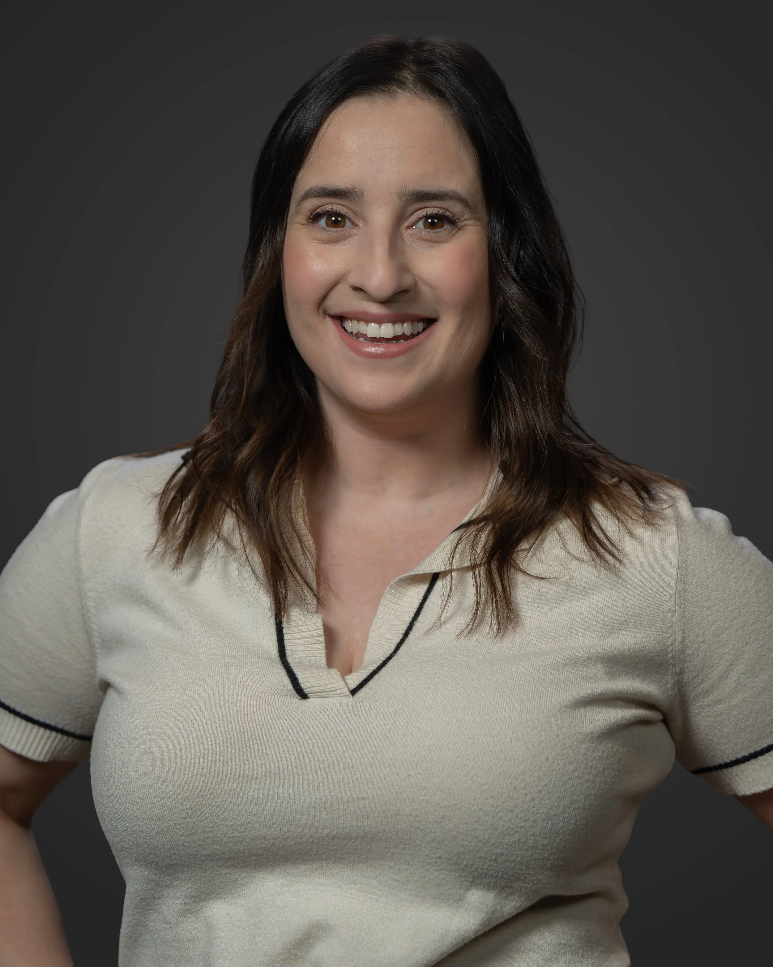 Portrait of a woman with shoulder-length brown hair, smiling, wearing a beige shirt with black trim, against a dark grey background.