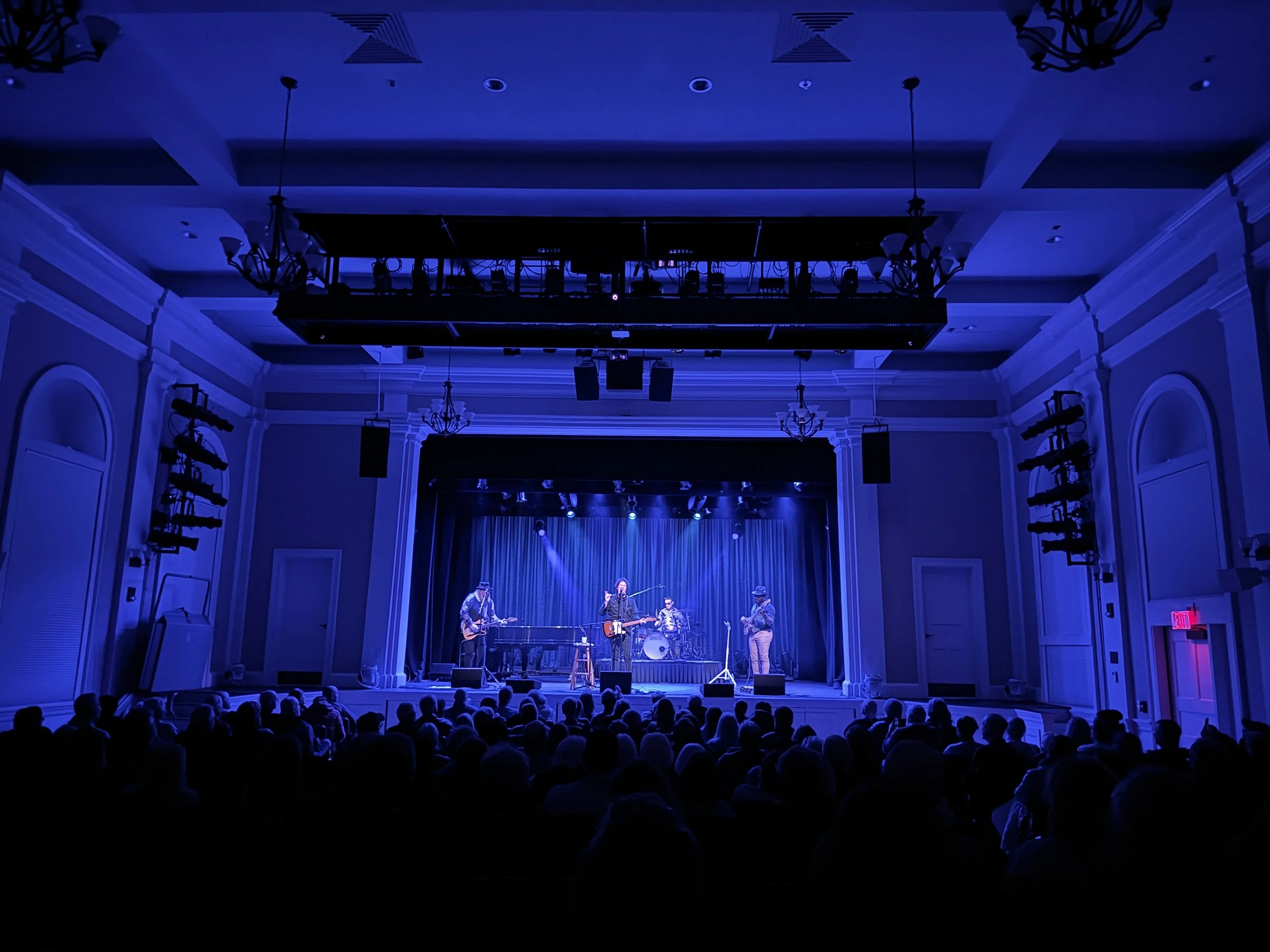Musicians performing on stage in a concert hall with blue lighting, audience seated in front, with chandeliers and stage equipment visible. live at The Kate in Old Saybrook, CT. Chrome Horse:The Bob Dylan Tribute