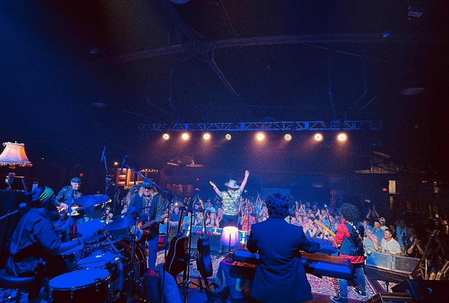 A live performance at Orange Peel. band on stage with a singer in a cowboy hat and zebra-print shirt engaging with the audience, who are dancing and enjoying the show. bright lights, and musical instruments, including drums and guitars, are visible.