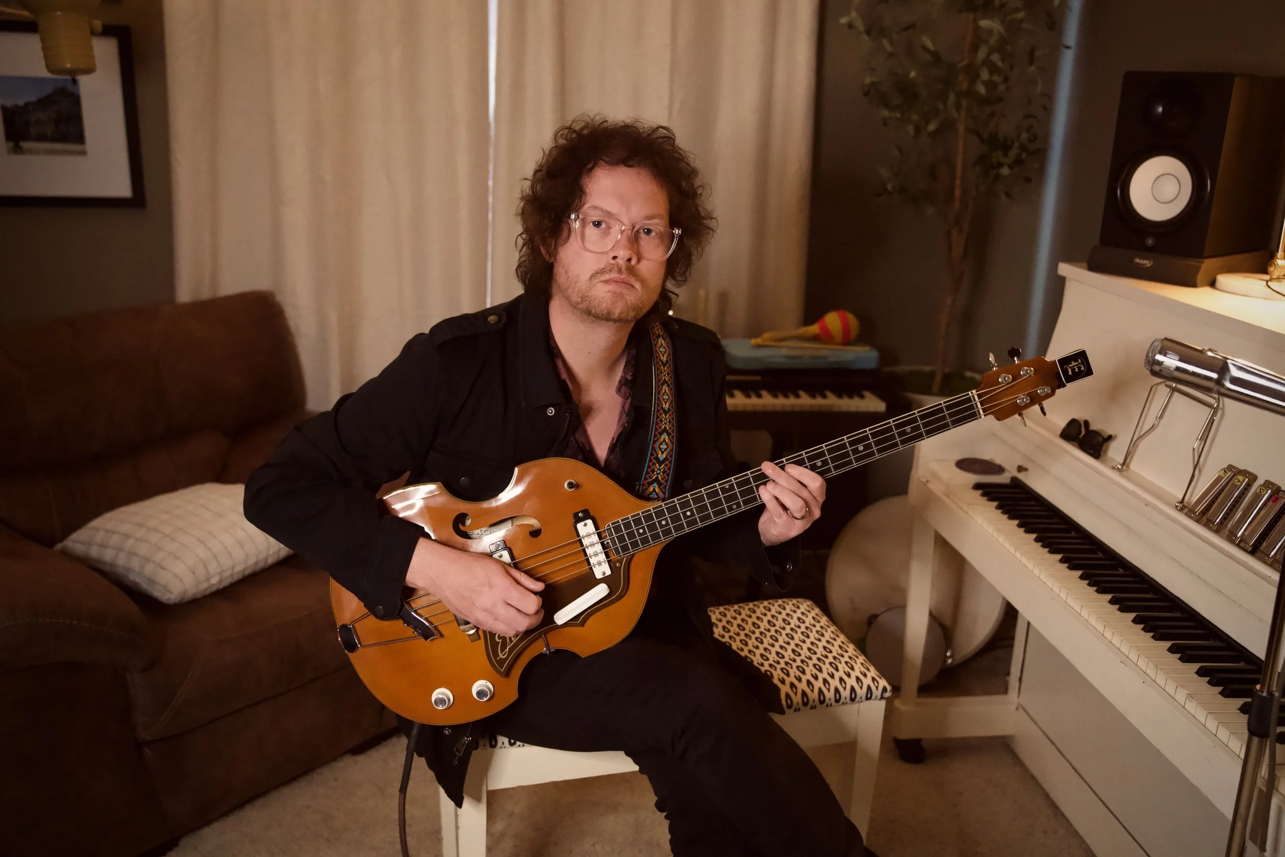 A man with curly hair and glasses sitting on a white chair, playing an electric guitar, in a cozy indoor setting with a brown sofa, a piano, and musical equipment. bass player