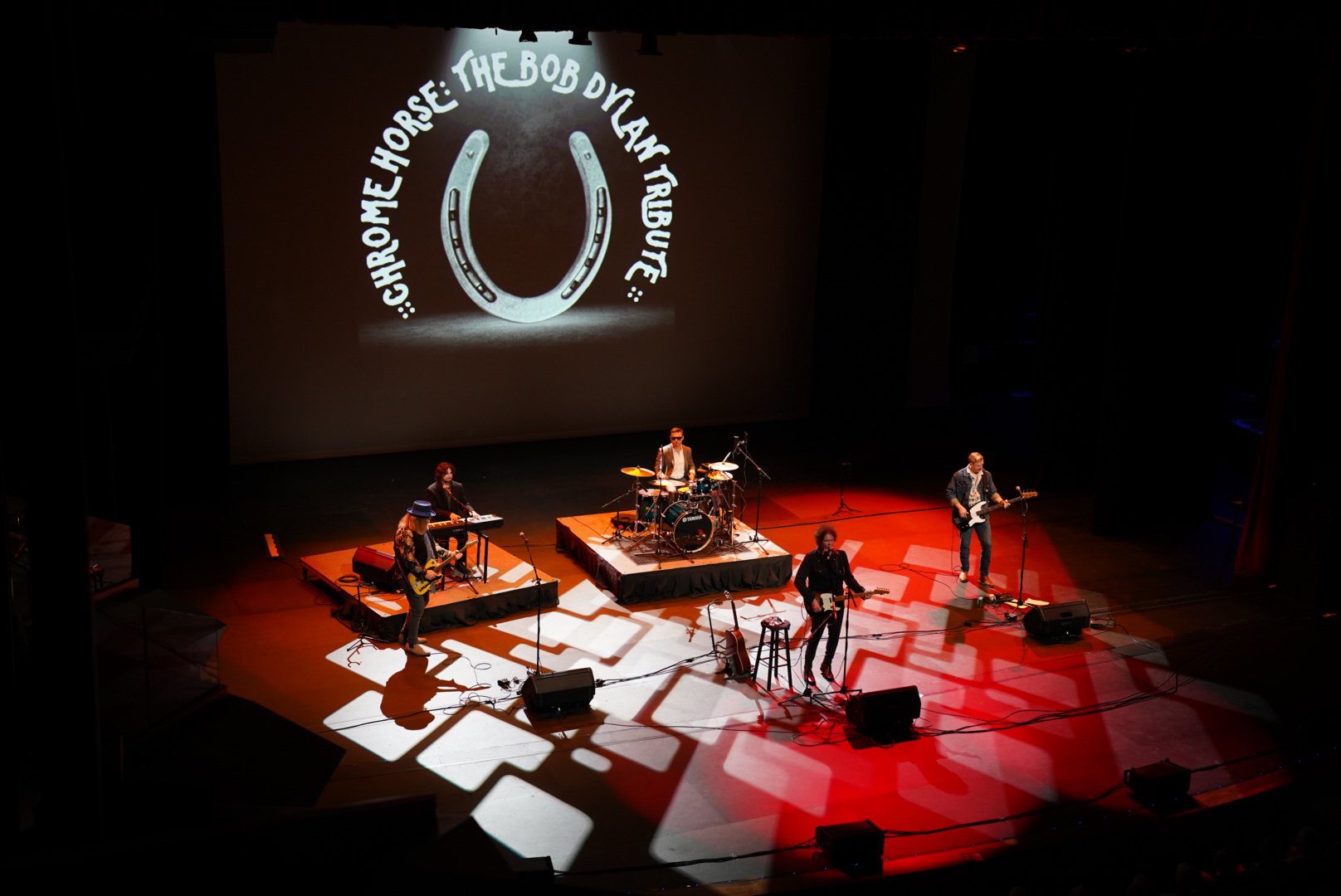 A band performing on stage with four musicians, one on drums, one on keyboard, and two on guitars. A large screen behind them displays the text 'Chrome Horse: The Bob Dylan Tribute' along with a horseshoe graphic. 