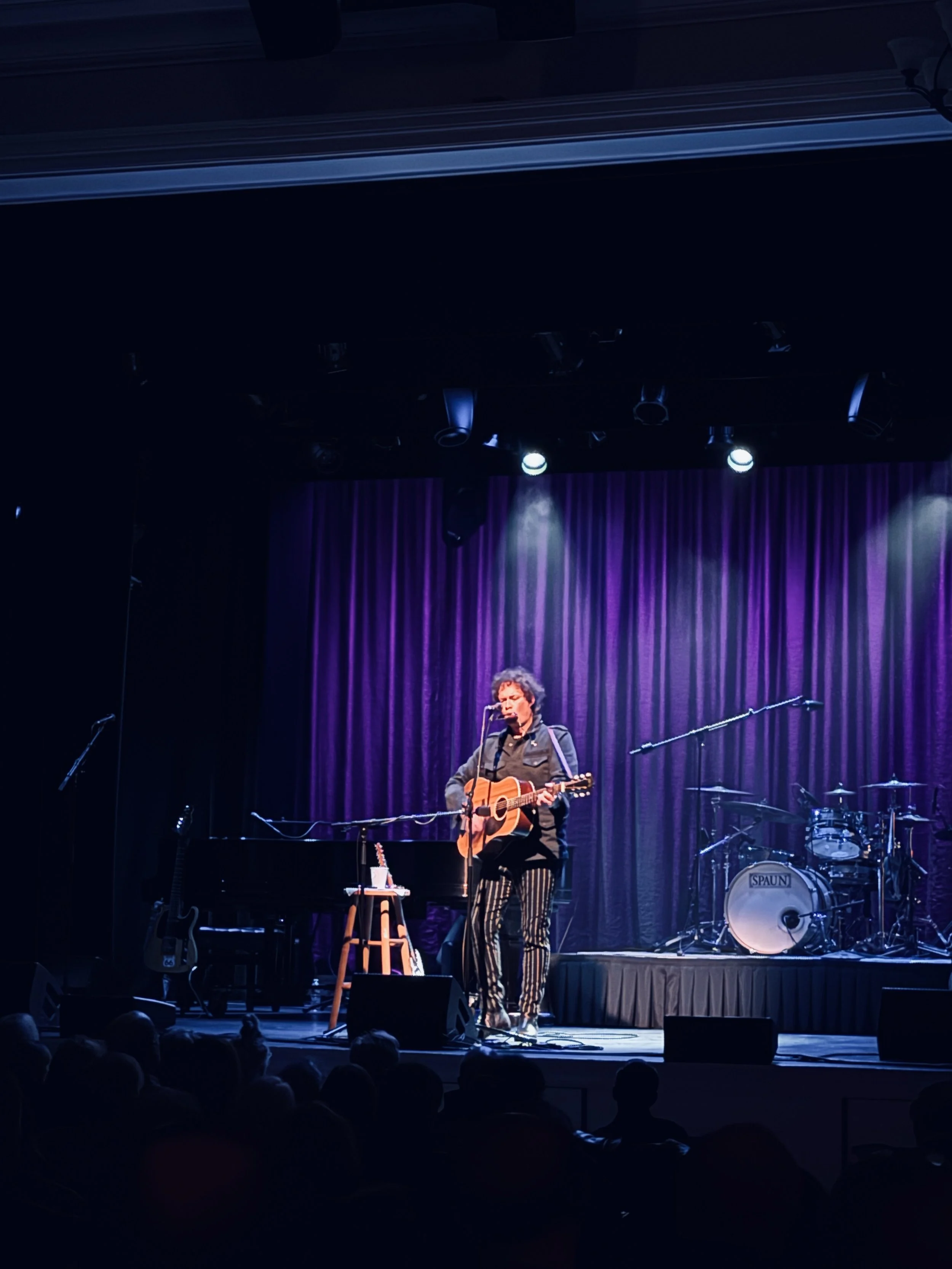 A musician on stage playing an acoustic guitar and singing into a microphone with purple curtains in the background