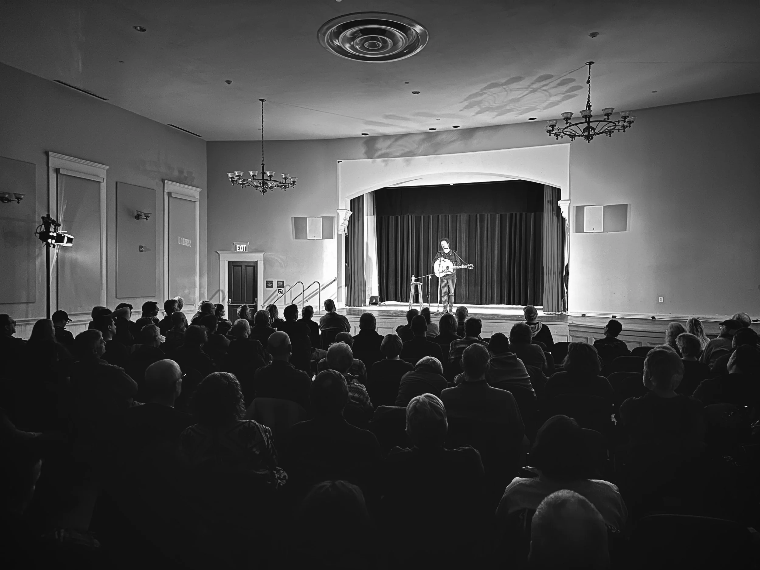 A black and white photo of a live music performance on a stage with a musician playing guitar and singing, facing an audience seated in a large room with chandeliers and wall sconces. Chrome Horse: The Bob Dylan Tribute. Chelmsford, Mass.